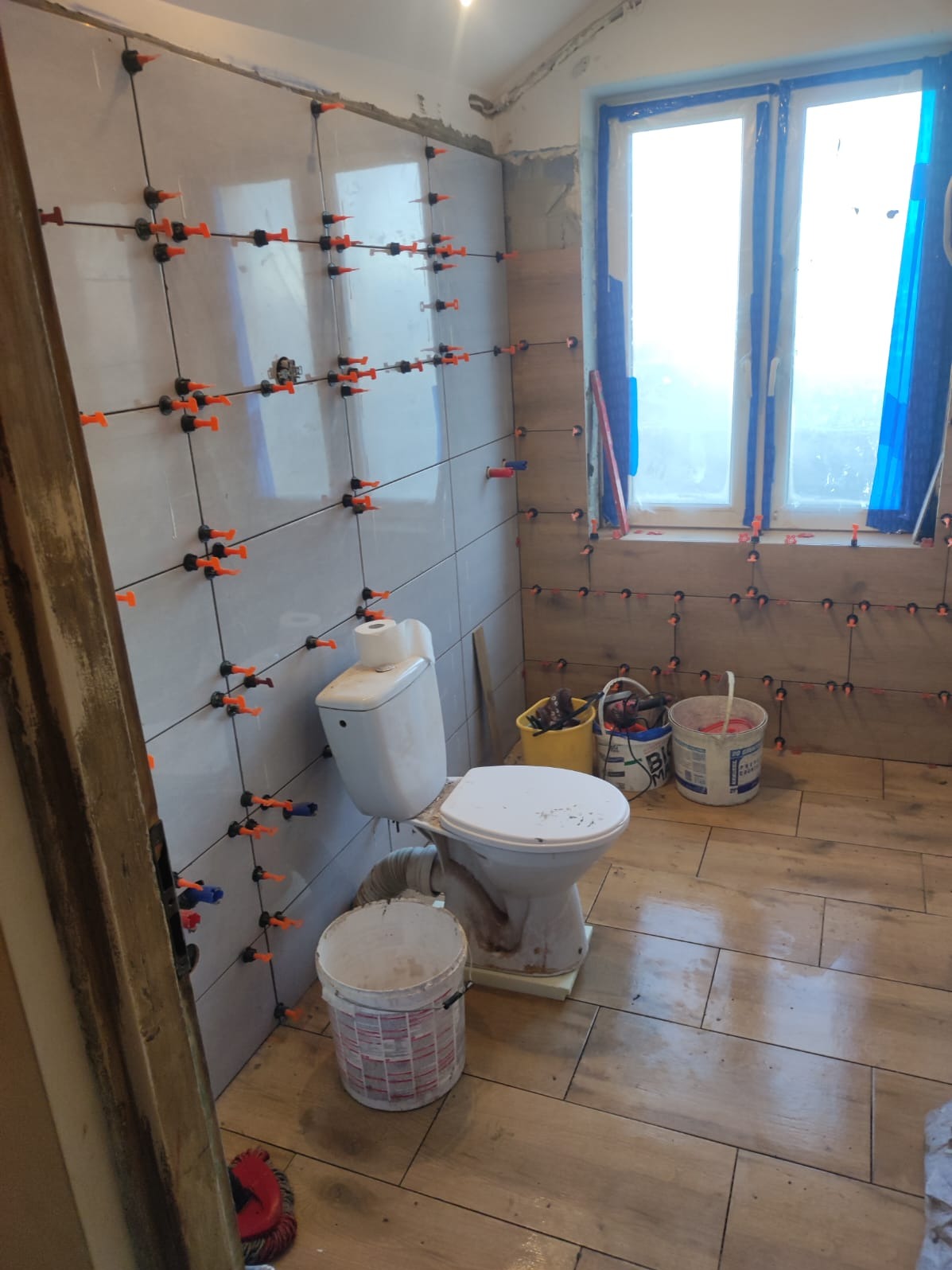 Układanie płytek w łazience z użyciem systemu poziomowania, widoczne narzędzia i fragment toalety. Prace wykończeniowe w trakcie realizacji.