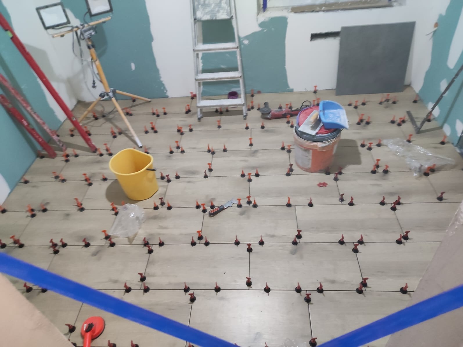 Układanie płytek podłogowych z użyciem systemu poziomowania. Widoczne narzędzia, drabina i lampa oświetleniowa. Ściany częściowo przygotowane do malowania.