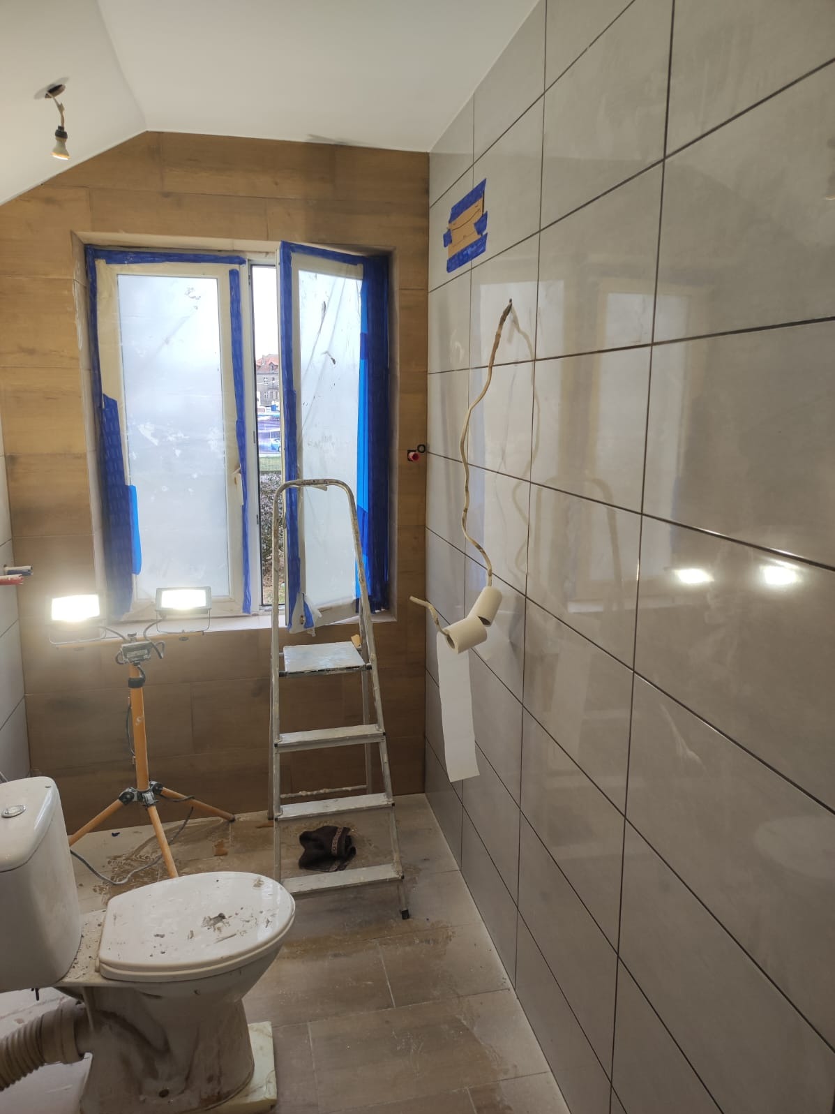 Remont łazienki: ułożone płytki na ścianach, zabezpieczone okno folią, drabina, lampa budowlana. Widoczne przewody elektryczne i niedokończone prace.