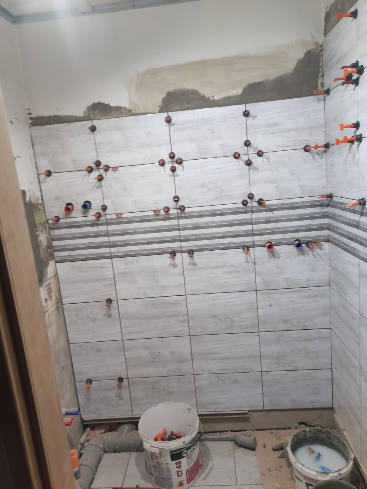 Układanie płytek ściennych w łazience, widoczne narzędzia glazurnicze i poziomice. Płytki imitujące drewno z szarym paskiem dekoracyjnym.