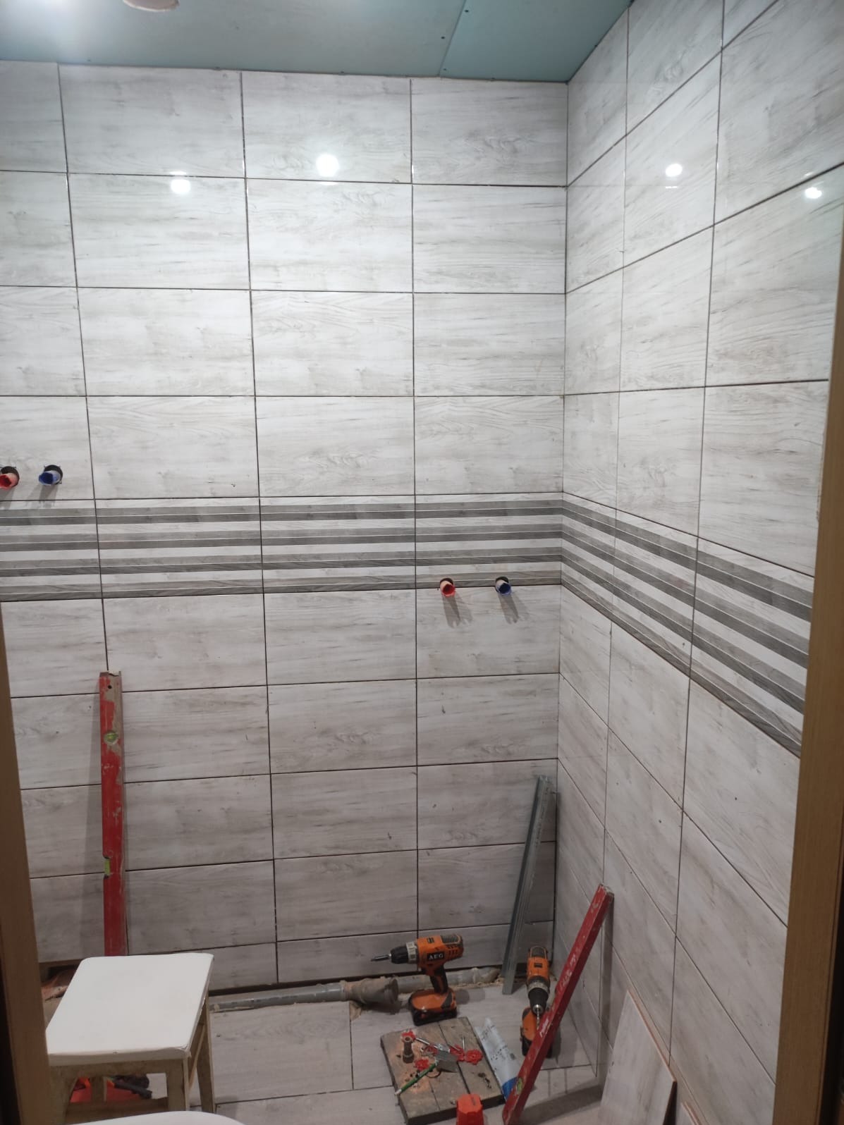 Wnętrze łazienki w trakcie wykańczania: jasne płytki imitujące drewno z dekoracyjnym pasem, widoczne narzędzia (wiertarki, poziomica) i instalacja wodna.