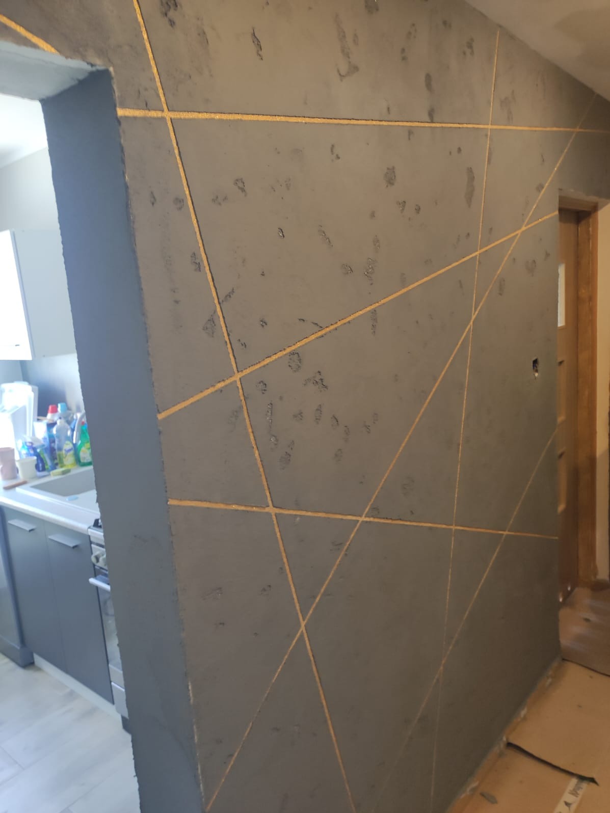 Szara ściana z dekoracyjnymi, złotymi liniami tworzącymi geometryczny wzór. Częściowo widoczny aneks kuchenny. Wykończenie wnętrza z efektem dekoracyjnym.