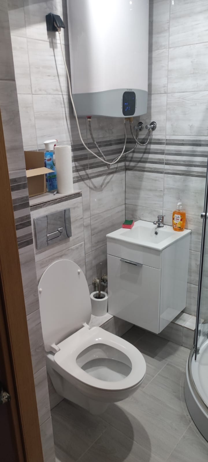 Wykończona łazienka w szarościach: podwieszana toaleta, biała szafka z umywalką, bojler i kabina prysznicowa. Widoczne akcesoria łazienkowe.