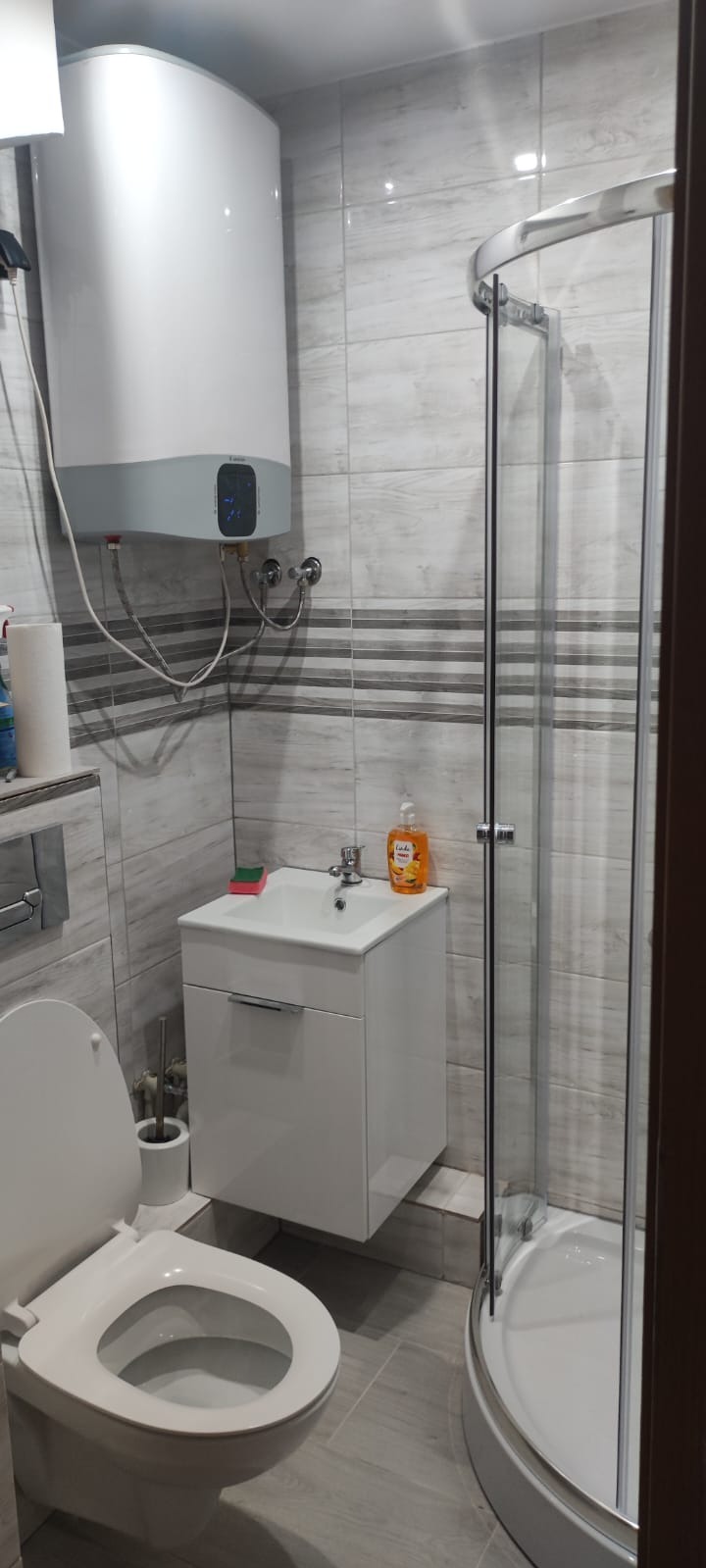 Jasna łazienka z kabiną prysznicową, umywalką z szafką i podgrzewaczem wody. Płytki w odcieniach szarości, minimalistyczny design. Widok na fragment toalety.