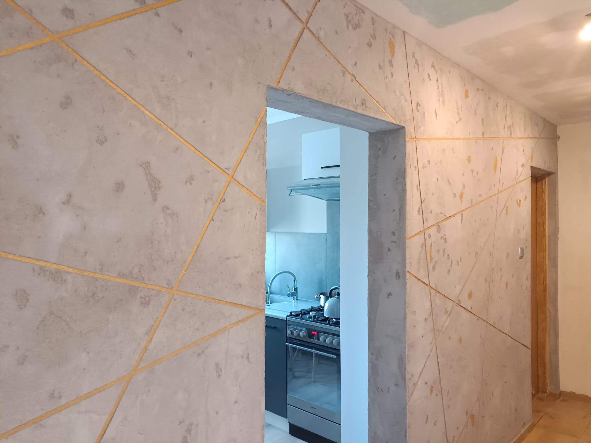 Ściana z efektem betonu architektonicznego i złotymi liniami, widok na kuchnię przez otwór drzwiowy. Wykończenie wnętrza w stylu nowoczesnym.