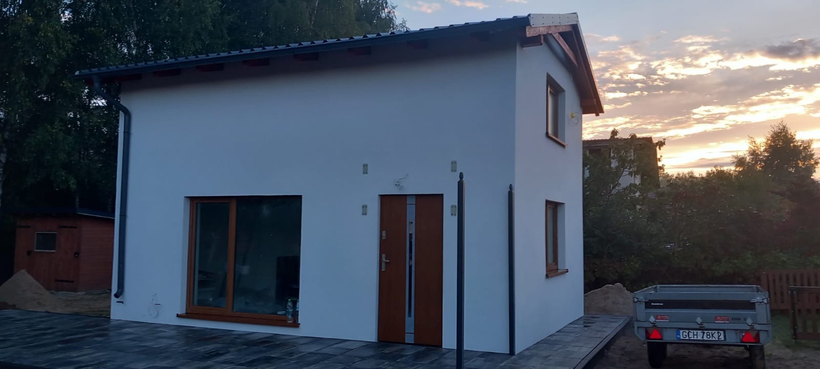 Biały, dwukondygnacyjny dom z brązowymi oknami i drzwiami, świeżo po elewacji, z widoczną kostką brukową i przyczepką w tle, sfotografowany o zmierzchu.