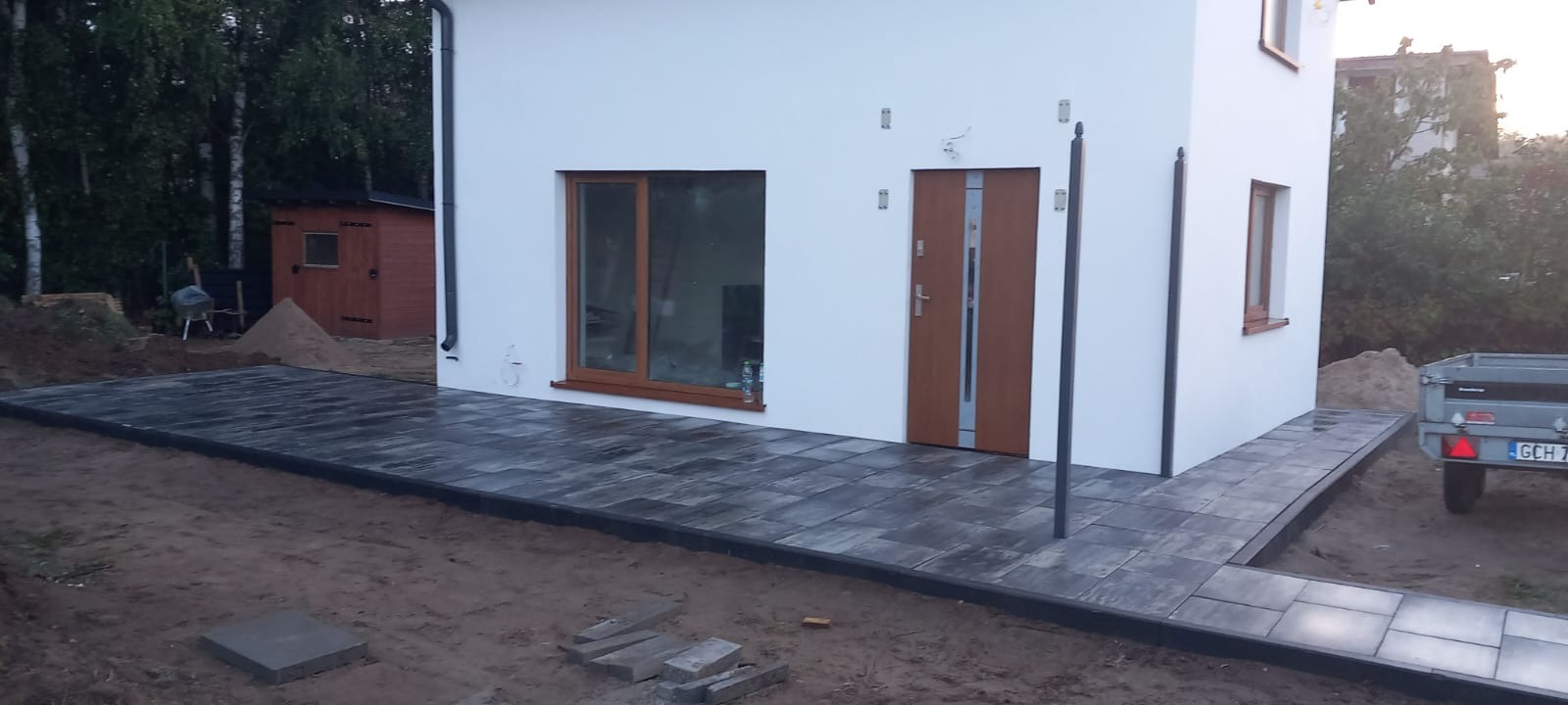 Nowoczesny dom z jasną elewacją i wybrukowanym tarasem z szarej kostki. Widoczny fragment przyczepy i drewniany budynek w tle.