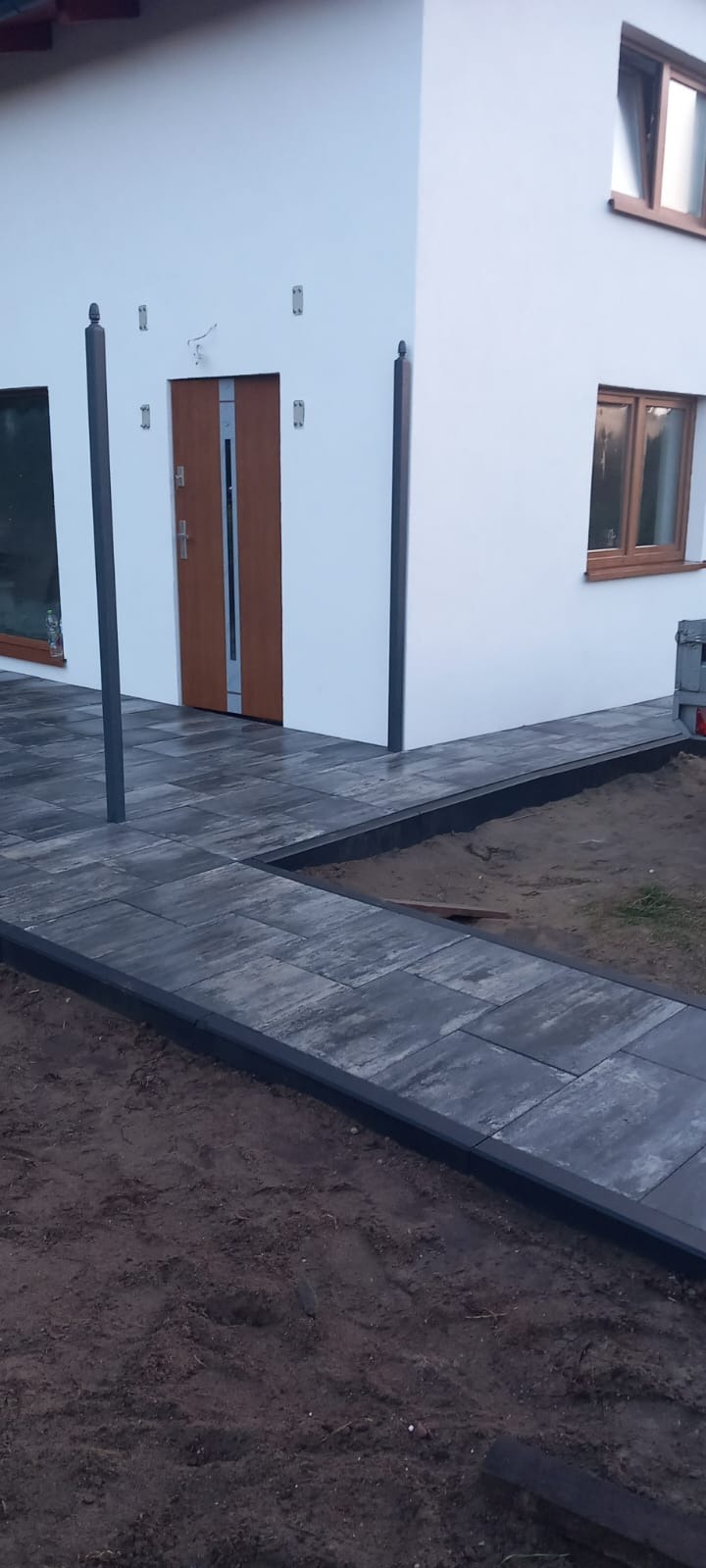 Nowoczesny dom z białymi ścianami i brukowanym podejściem z szarych płyt. Widoczne okna z drewnianą ramą i brązowe drzwi wejściowe.