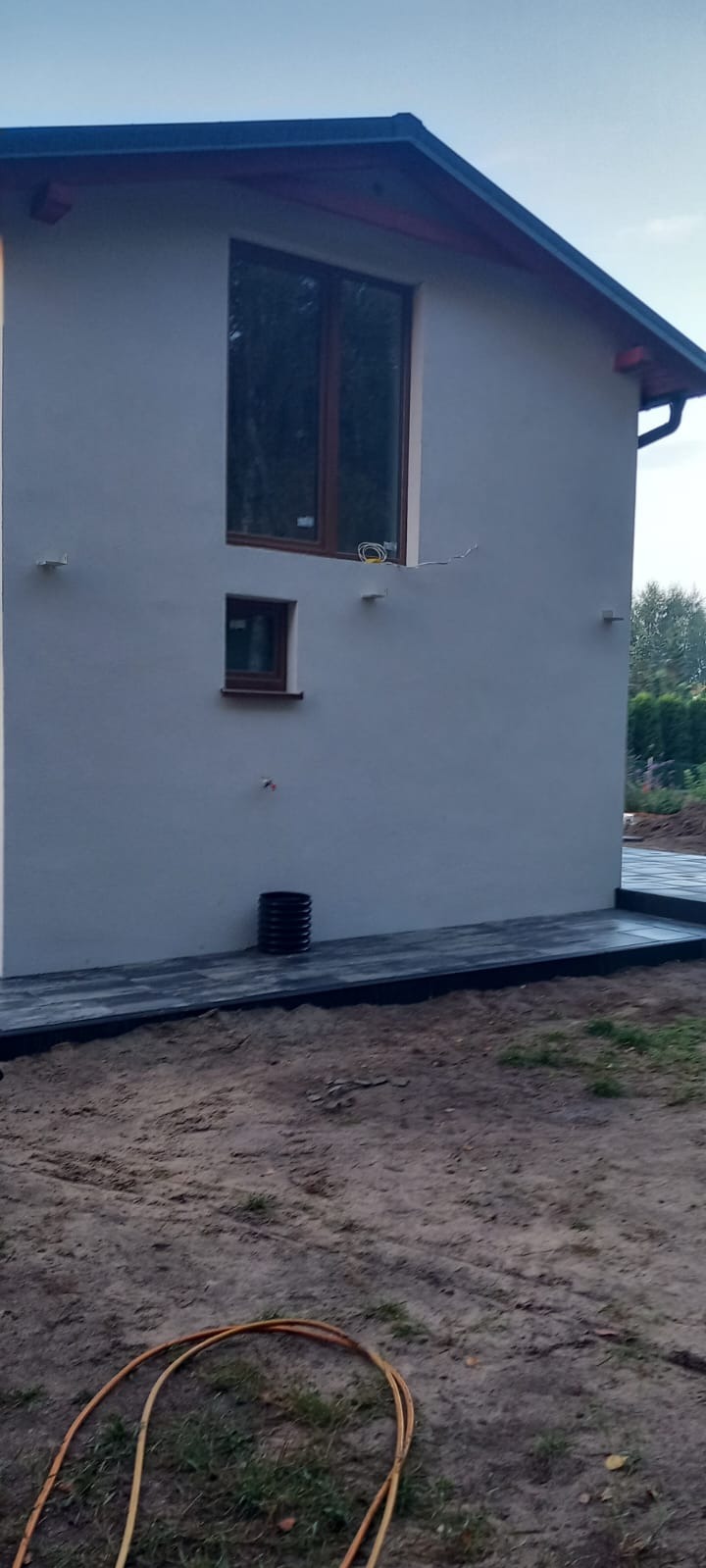 Wykończona elewacja domu z dwoma oknami i czarną rurą spustową przy tarasie z kostki. Widoczne przewody elektryczne przy górnym oknie.
