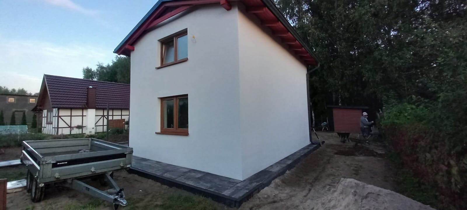 Biała elewacja domu z brązowymi oknami i czerwonym dachem, widok z boku na tle zieleni, przyczepa i kostka brukowa w otoczeniu.