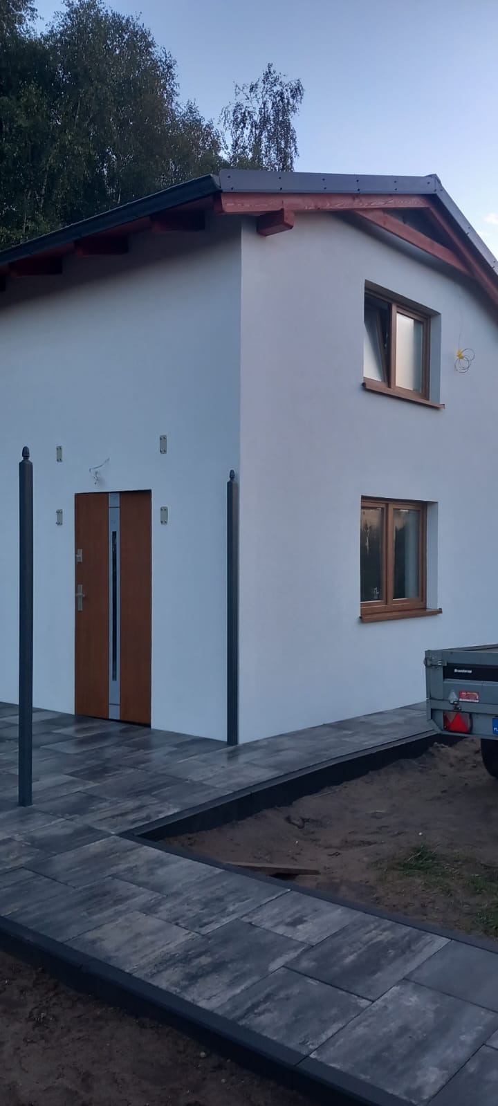 Biała elewacja domu jednorodzinnego z drewnianą stolarką okienną i brukowanym chodnikiem w Chojnicach. Widoczne wykończone detale budynku.