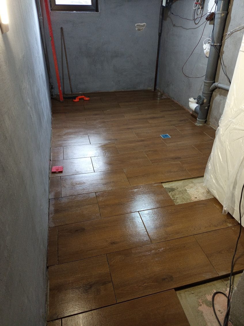 Układanie brązowych płytek imitujących drewno w pomieszczeniu z szarą ścianą. Widoczny fragment podłogi bez płytek i narzędzia glazurnicze.