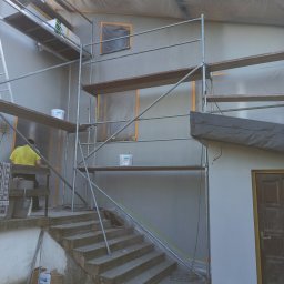 M&M HOUSE - Elewacja domu w trakcie tynkowania, widoczne rusztowania, wiaderka z tynkiem i zabezpieczone okna folią. Pracownik w żółtej koszulce na rusztowaniu.