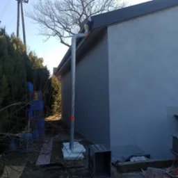 Szara elewacja budynku z orynnowaniem, widok z dołu. Na pierwszym planie skrzynki i elementy budowlane. W tle drzewa i słupy energetyczne.