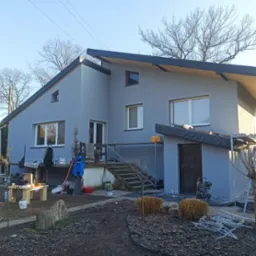 Szara elewacja domu jednorodzinnego w Opolu po malowaniu. Widoczne narzędzia i materiały malarskie. Uwagę zwraca nowoczesna bryła budynku i minimalistyczny styl.