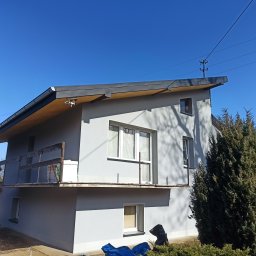 M&M HOUSE - Dom z szarą elewacją w słoneczny dzień. Widoczny balkon z drewnianą barierką i fragment elewacji z oknami. Częściowo zakryty materiał budowlany.