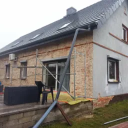 Dom w trakcie renowacji elewacji: fragment ściany z odsłoniętą czerwoną cegłą, obok otynkowana szara ściana, rusztowania, okna z roletami.