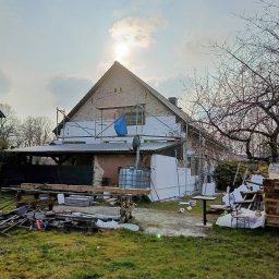 M&M HOUSE - Dom w trakcie ocieplania: biały styropian na elewacji, rusztowania, częściowo otynkowana ściana szczytowa, materiały budowlane na trawniku. Widok w słoneczny dzień.