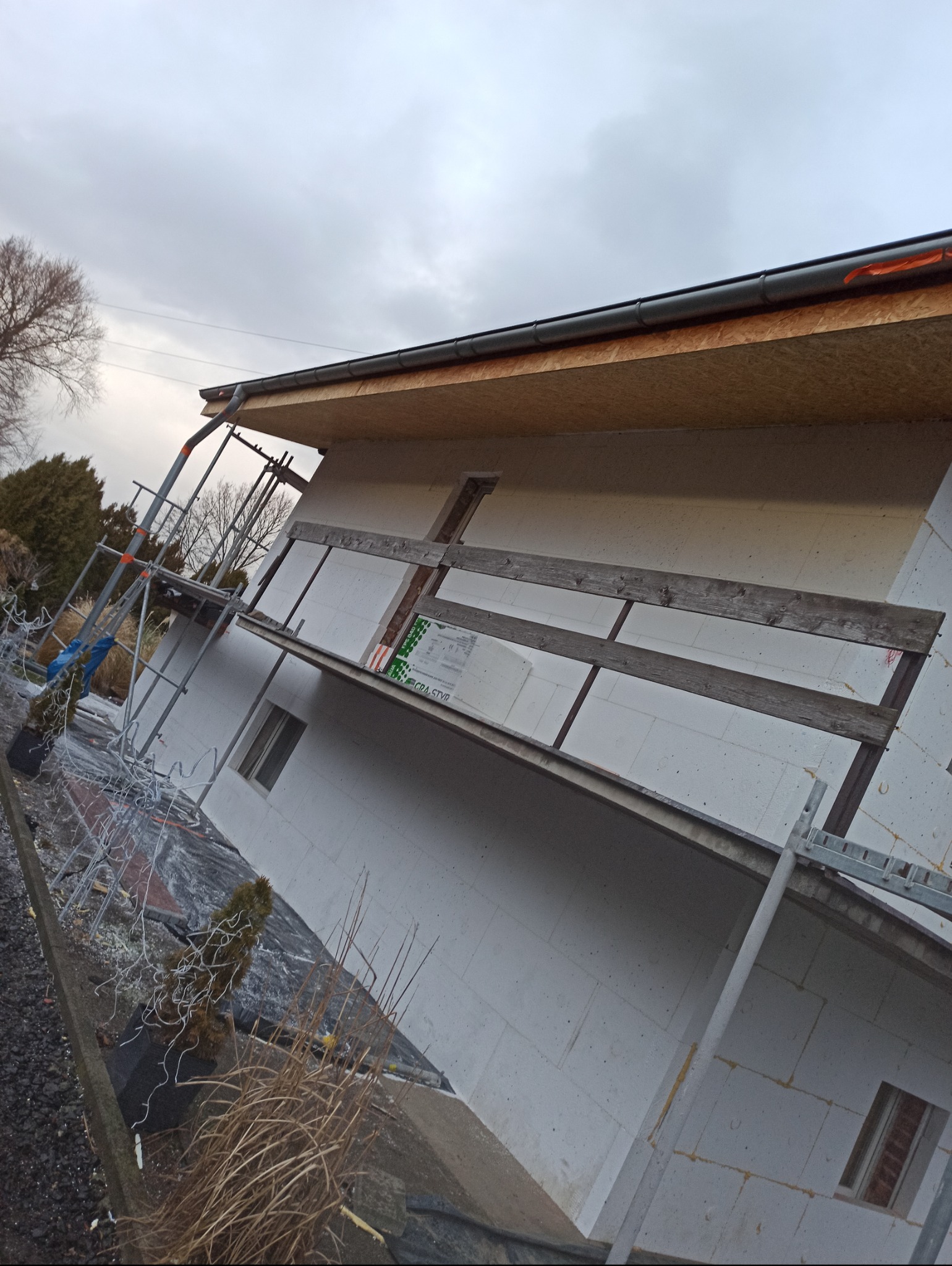 Budowa elewacji domu jednorodzinnego z białych bloczków, widoczne rusztowania i materiały izolacyjne, pochmurne niebo w tle. Detal budowlany w trakcie prac.