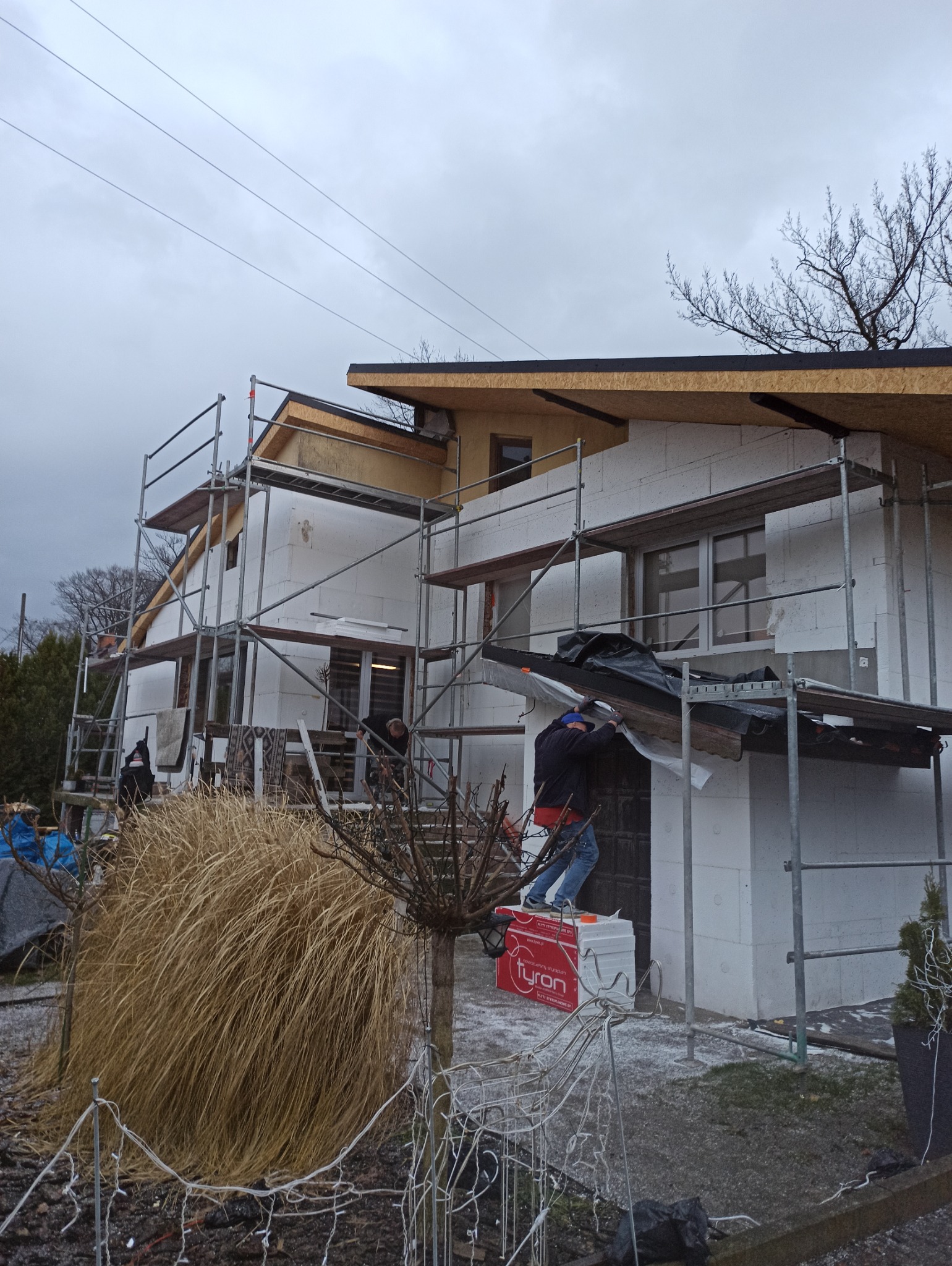 Dom w trakcie ocieplania: styropianowe płyty na ścianach, rusztowania, pracownicy montujący elewację. Widoczne materiały izolacyjne marki Tyron.