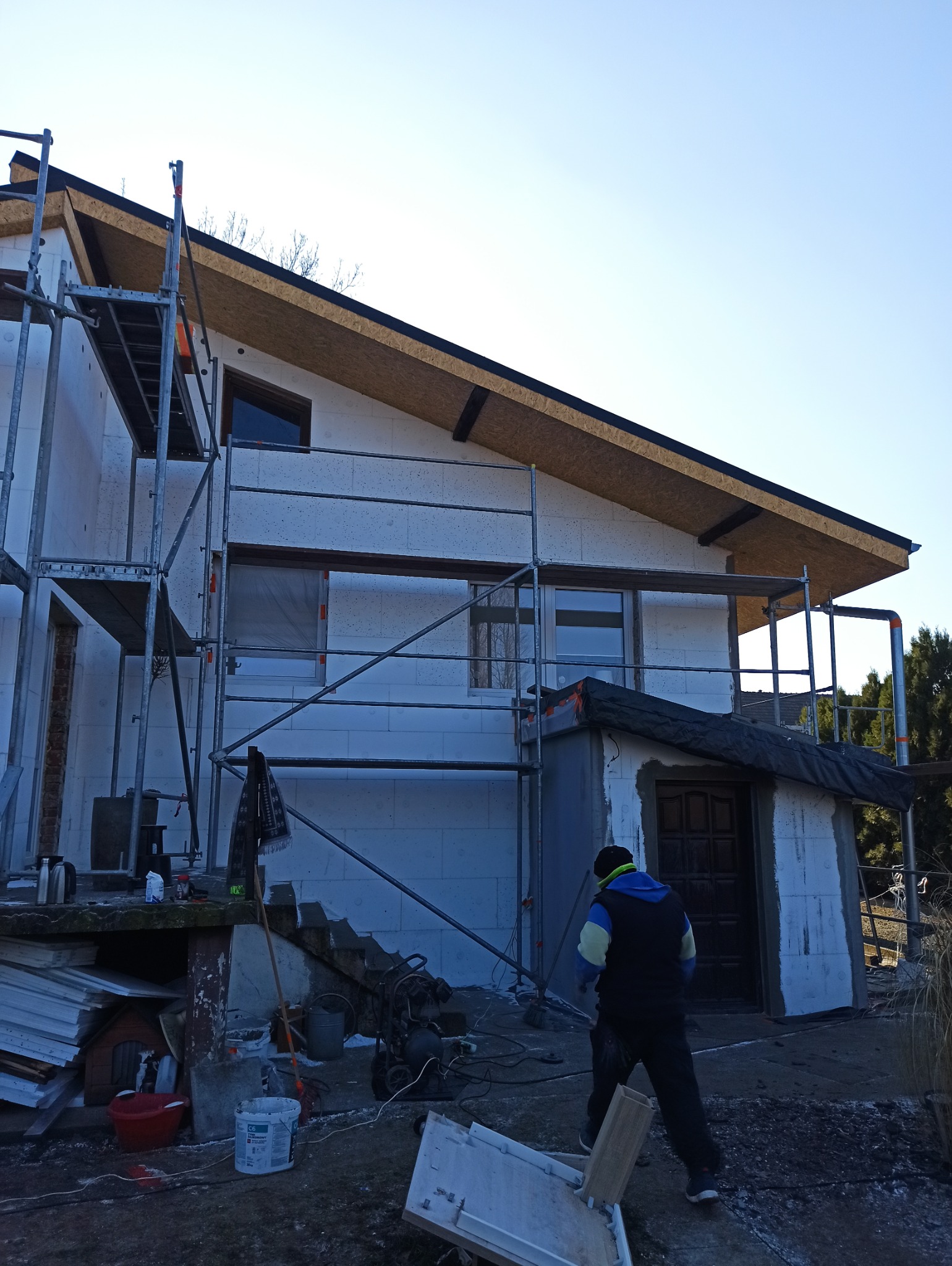 Dom w trakcie ocieplania, widoczne rusztowania i styropian na ścianach. Osoba w roboczym ubraniu w pobliżu. Ujęcie z dołu, podkreślające wysokość budynku.