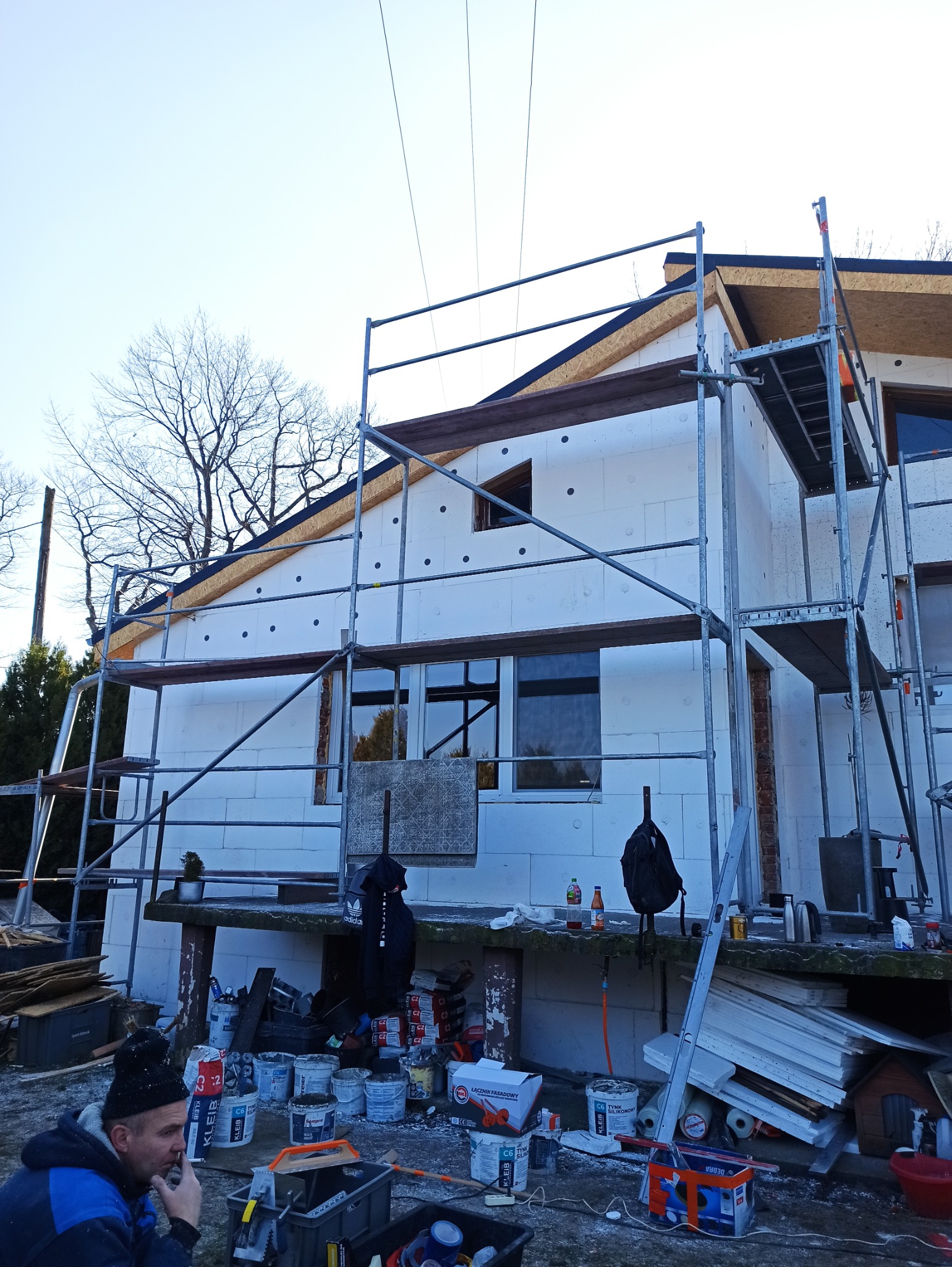 Ocieplanie domu styropianem w Opolu. Widoczne rusztowania, materiały budowlane i pracownik podczas przerwy. Etap przed tynkowaniem.