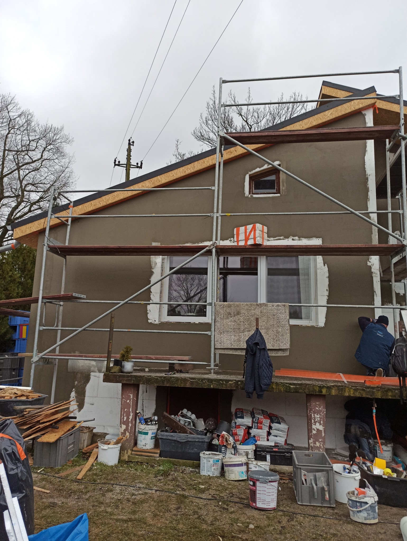 Dom w trakcie tynkowania elewacji, widoczne rusztowania, materiały budowlane i pracownik w trakcie nakładania tynku. Częściowo otynkowana ściana.