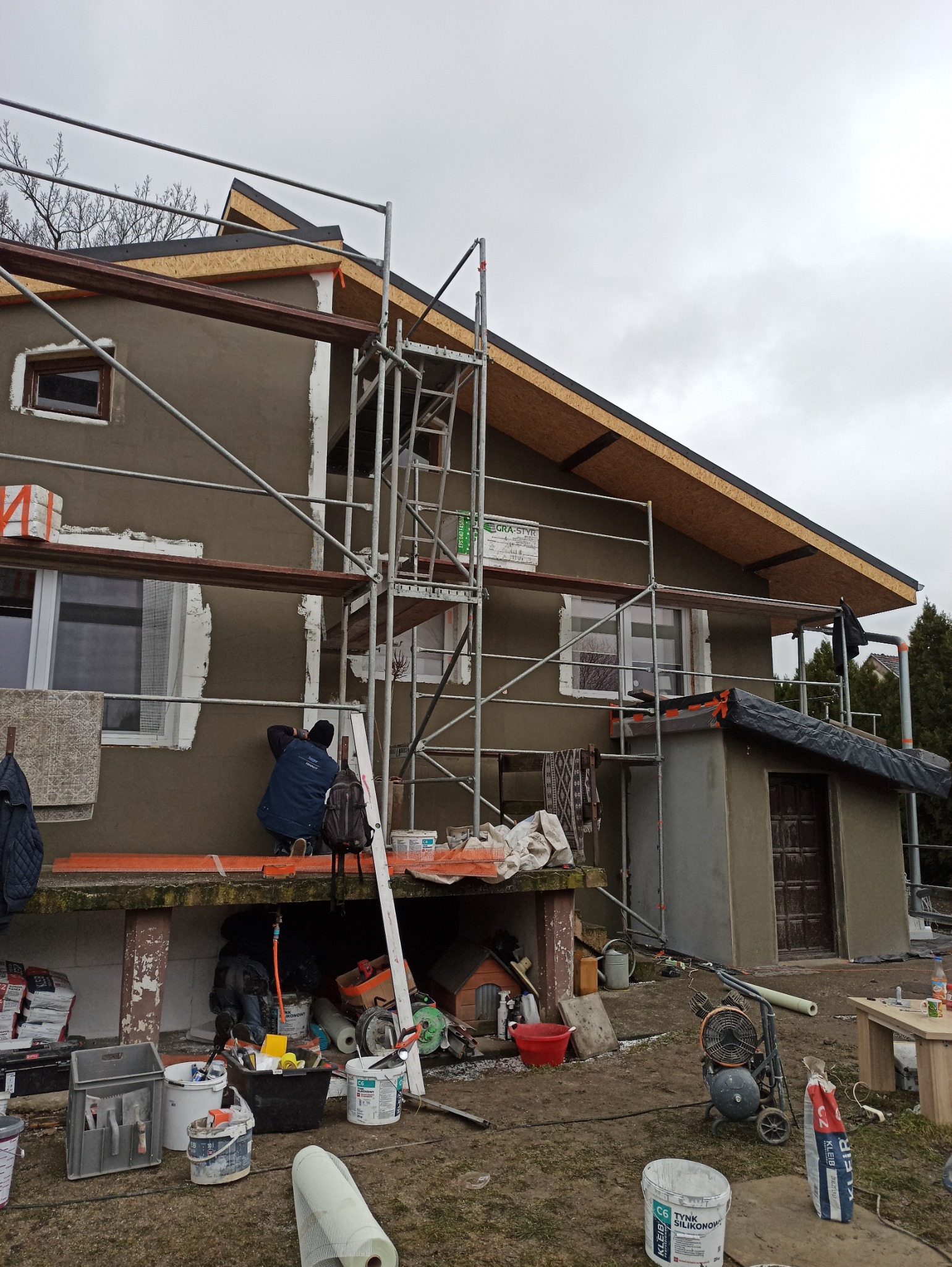 Prace tynkarskie na elewacji domu jednorodzinnego, widoczne rusztowania i narzędzia. Pracownik w trakcie nakładania tynku. Częściowo otynkowana ściana budynku.