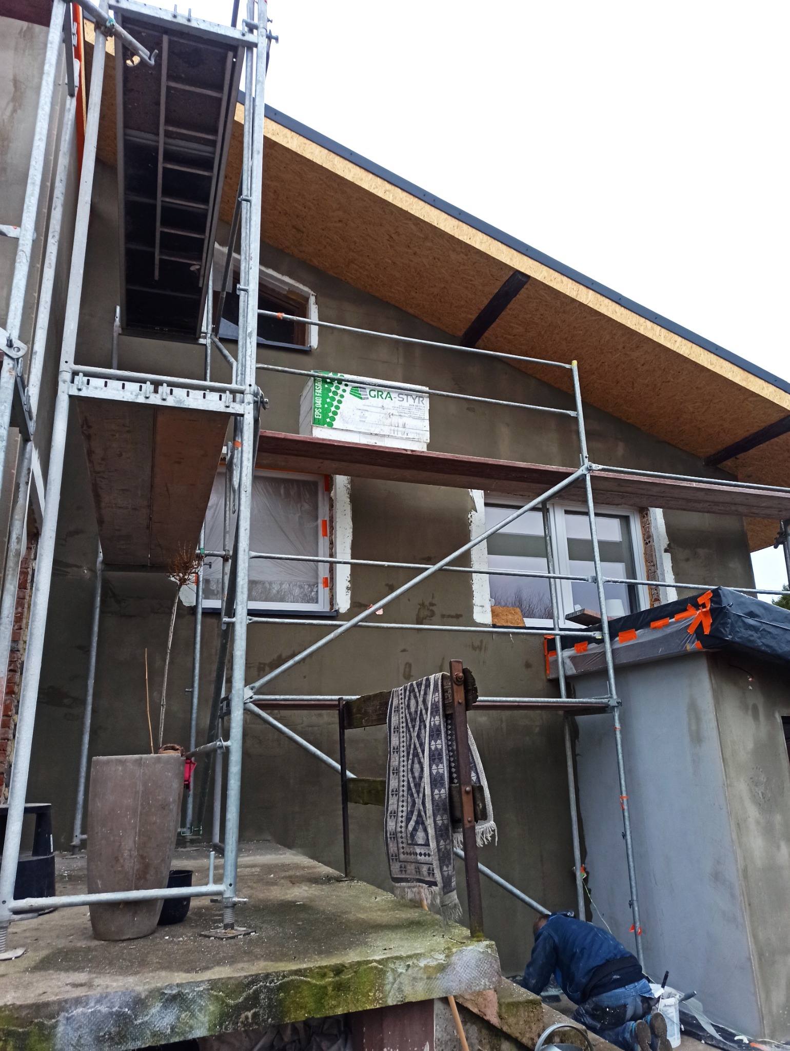 Dom w trakcie ocieplania: szare tynki, rusztowania, styropian GRA-STYR. Okna zabezpieczone folią. Na pierwszym planie dywan na barierce i pracownik na kolanach.
