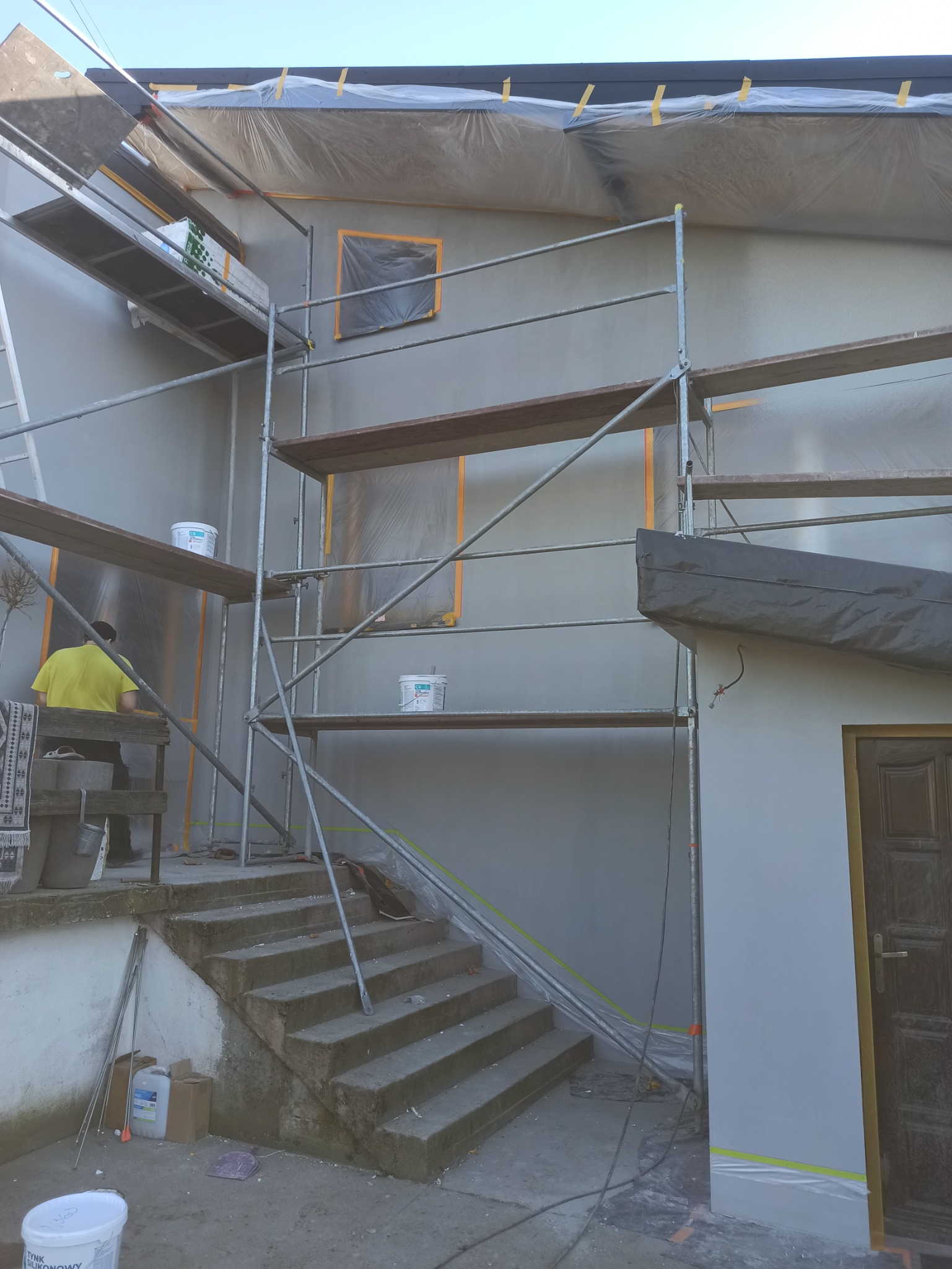 Elewacja domu w trakcie tynkowania, widoczne rusztowania, wiaderka z tynkiem i zabezpieczone okna folią. Pracownik w żółtej koszulce na rusztowaniu.