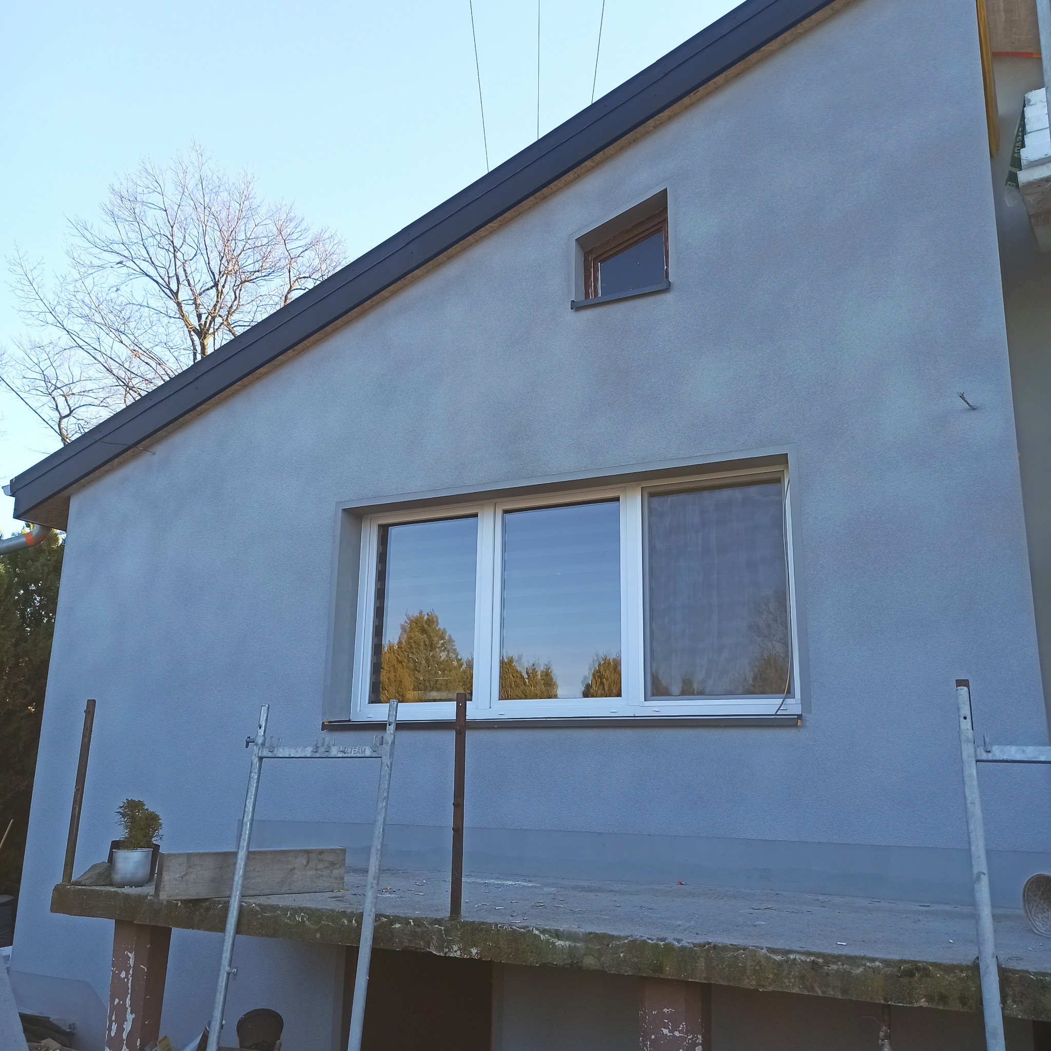 Świeżo otynkowana elewacja domu jednorodzinnego w odcieniach szarości, z widocznymi oknami i elementami rusztowania. Minimalistyczny design, gładka faktura tynku.