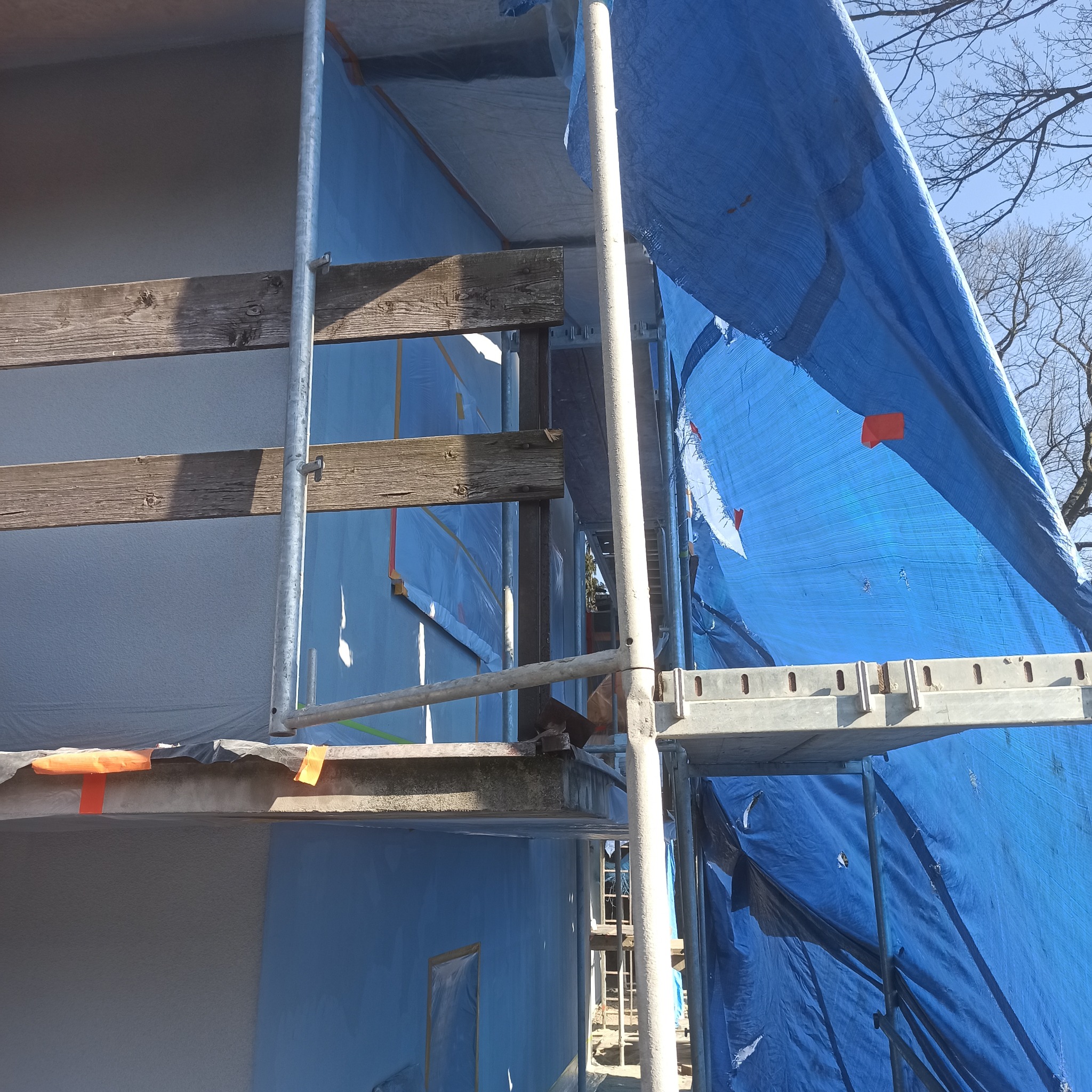 Elewacja budynku w trakcie prac, osłonięta niebieską plandeką i zabezpieczona rusztowaniem. Widoczne drewniane elementy konstrukcyjne i fragmenty tynku.