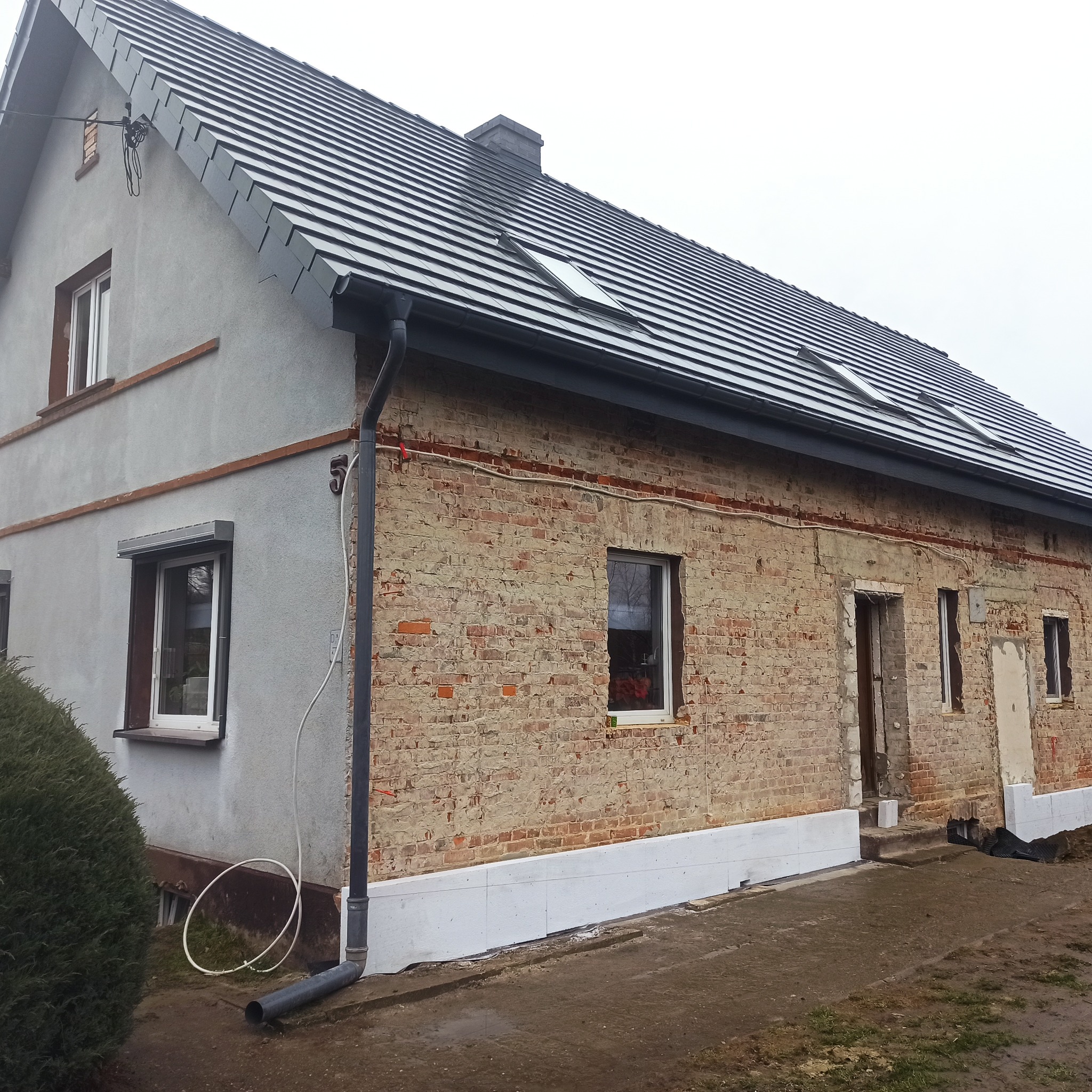 Dom z częściowo otynkowaną elewacją i odsłoniętą cegłą, widoczne okna dachowe i rynna. Część fundamentów ocieplona białym styropianem.
