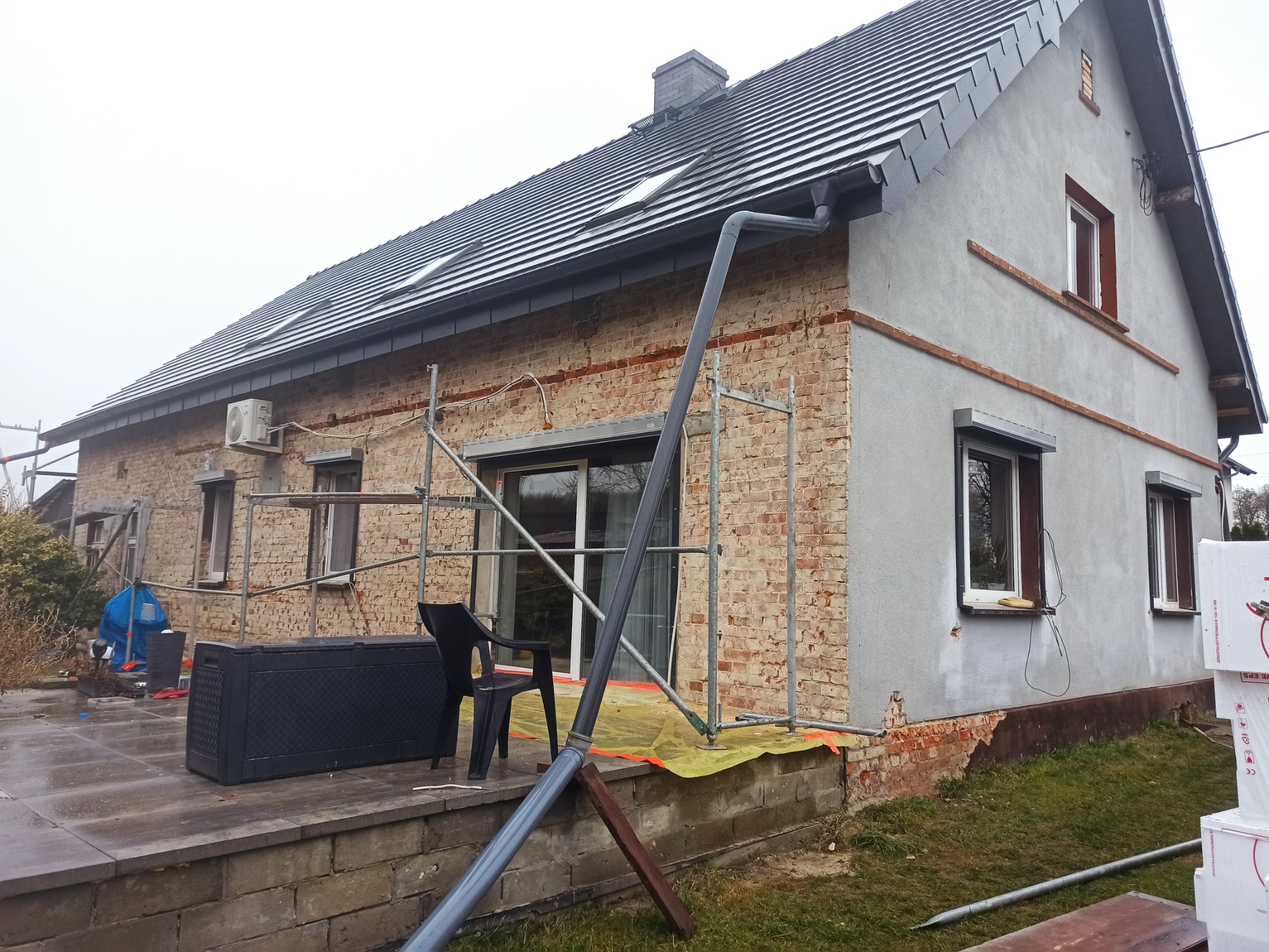 Dom w trakcie renowacji elewacji: fragment ściany z odsłoniętą czerwoną cegłą, obok otynkowana szara ściana, rusztowania, okna z roletami.