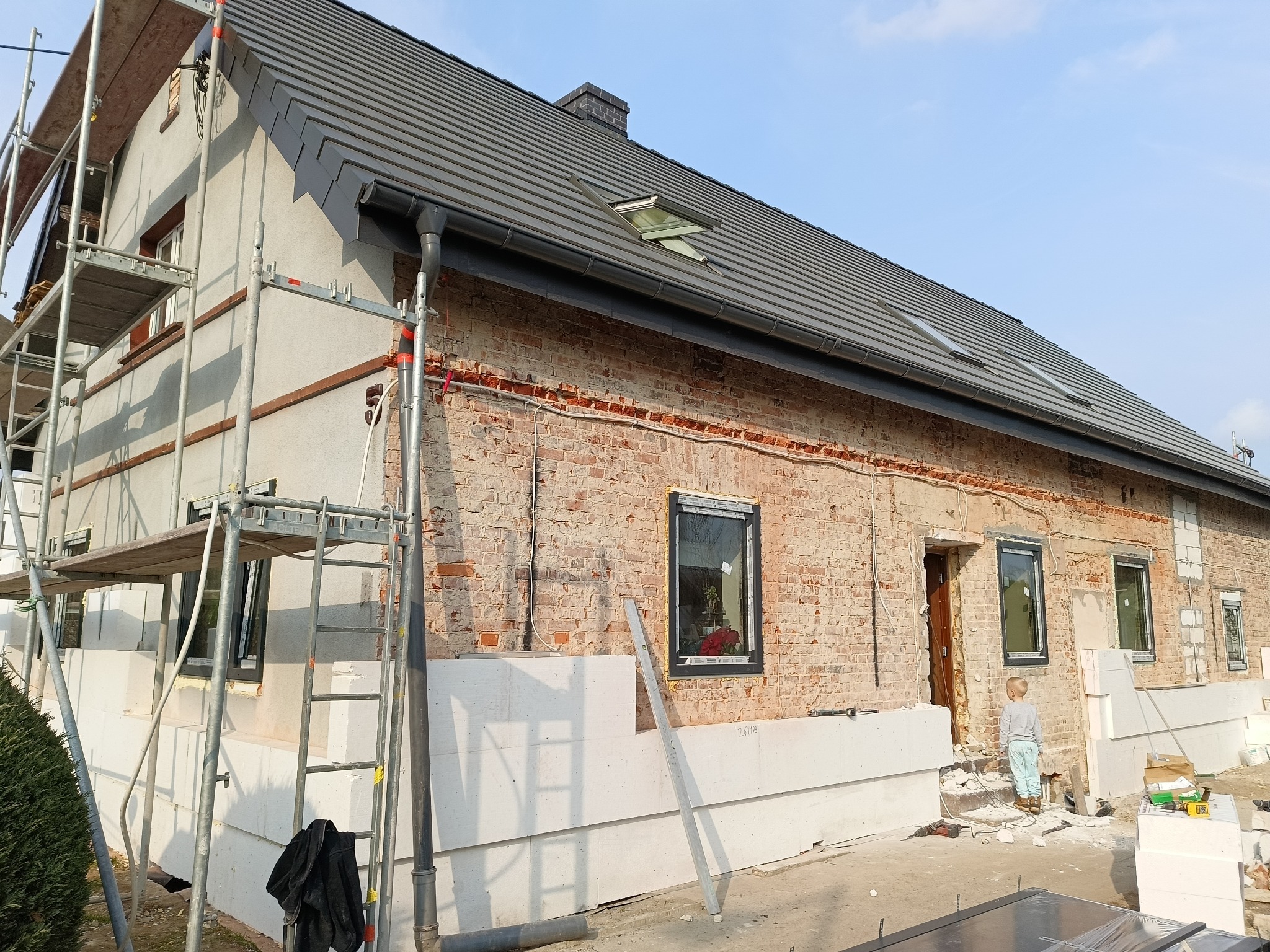 Dom w trakcie ocieplania: rusztowania, biały styropian na elewacji, fragment odsłoniętej cegły. Dziecko stoi przed wejściem do budynku.