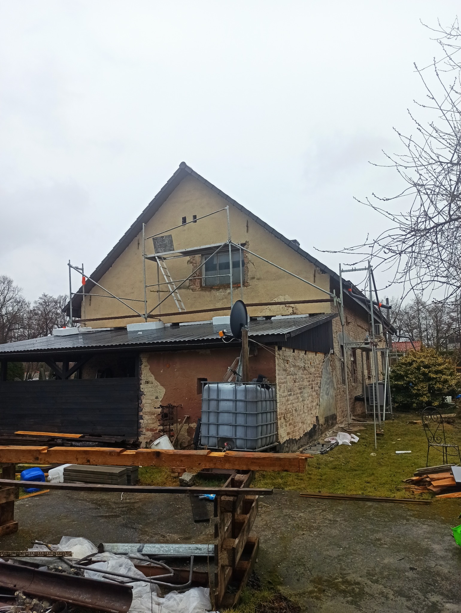 Dom w trakcie renowacji elewacji z widocznymi rusztowaniami i elementami przygotowawczymi, w pochmurny dzień. Widoczne uszkodzenia tynku i cegieł.