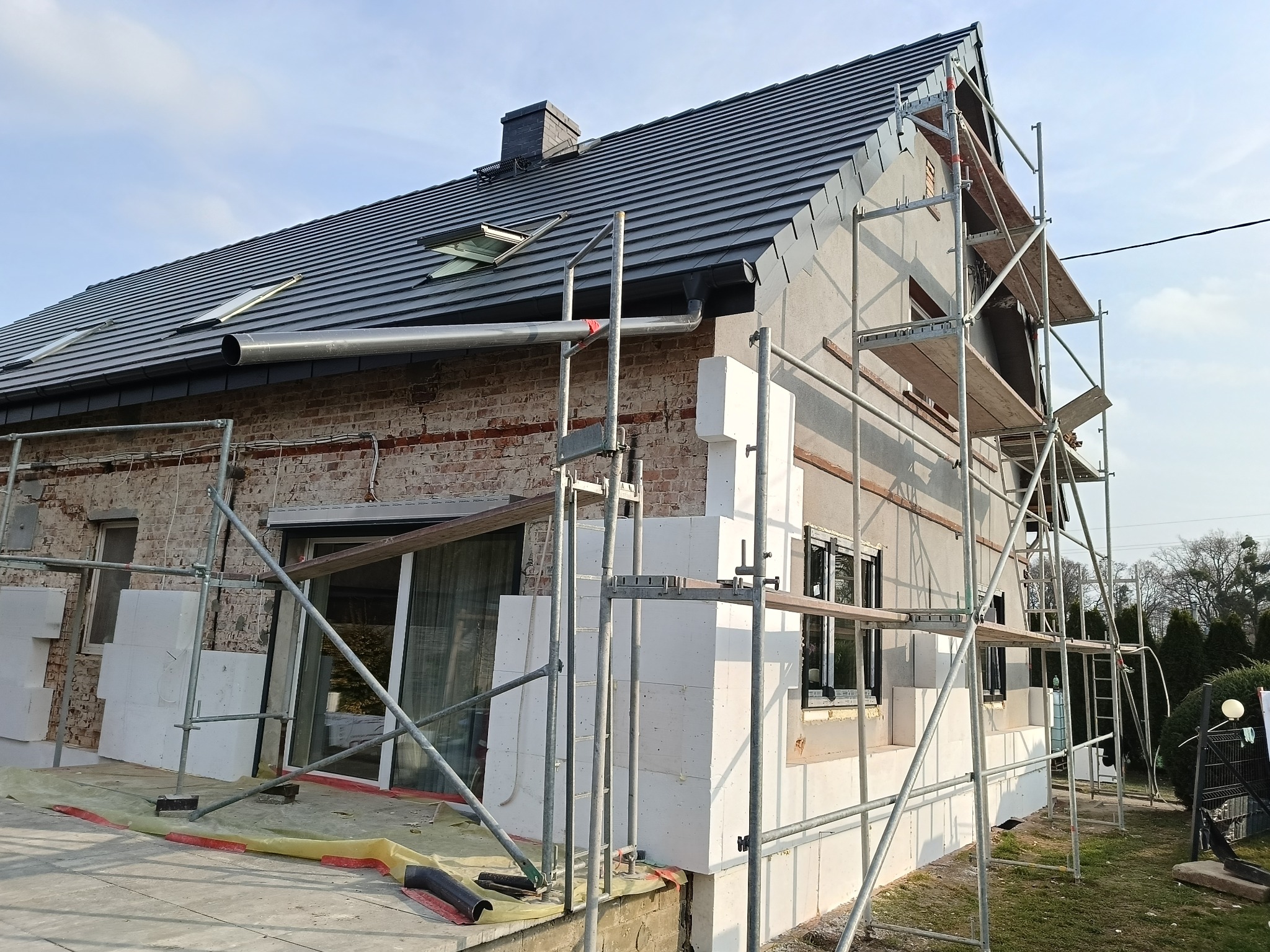 Dom w trakcie ocieplania: część ściany z cegły, część z ociepleniem, rusztowania. Widoczne okna, dach z dachówkami i rynna. Prace elewacyjne w toku.
