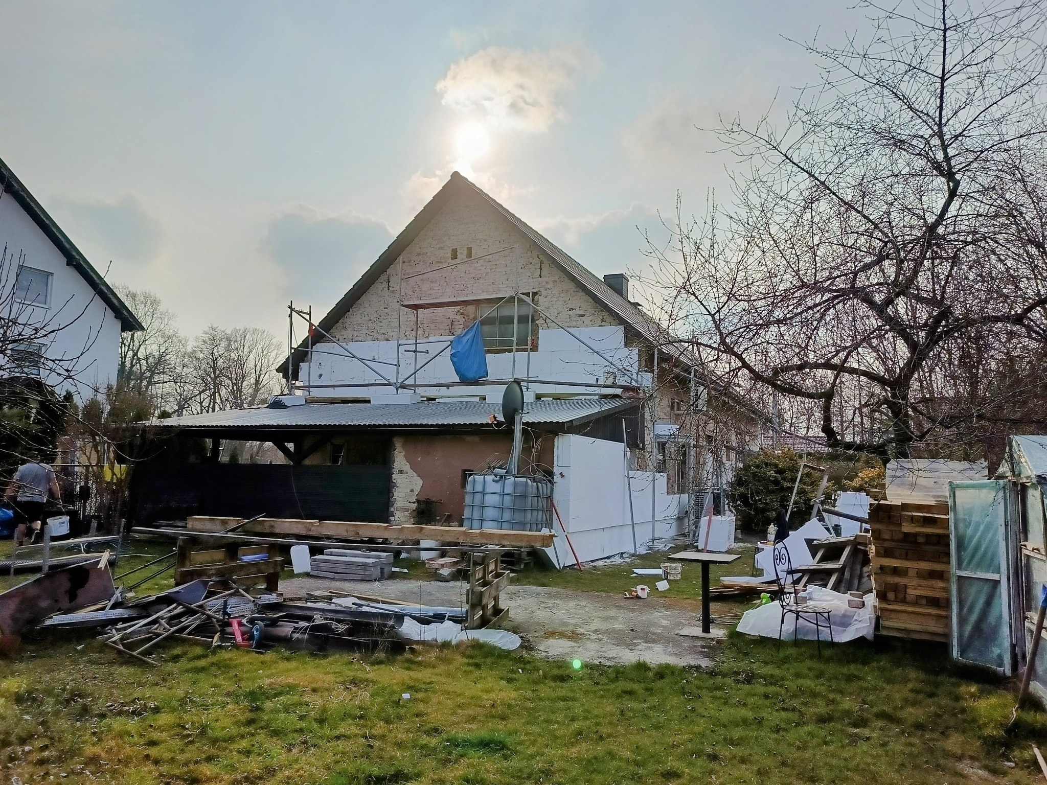 Dom w trakcie ocieplania: biały styropian na elewacji, rusztowania, częściowo otynkowana ściana szczytowa, materiały budowlane na trawniku. Widok w słoneczny dzień.