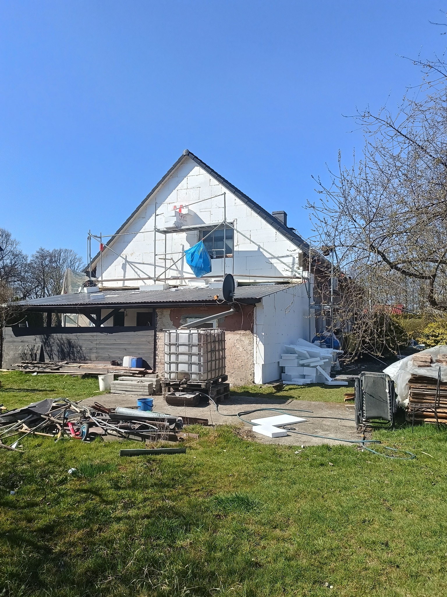 Dom w trakcie ocieplania: biały styropian na elewacji, rusztowania, niebieska plandeka. Widoczne materiały budowlane i narzędzia na trawniku. Słoneczny dzień.