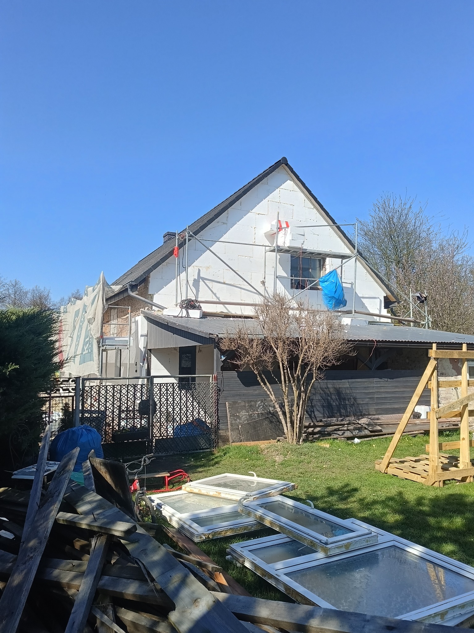 Dom w trakcie ocieplania elewacji, widoczne rusztowania i styropianowe płyty. Na pierwszym planie okna i drewniane elementy.