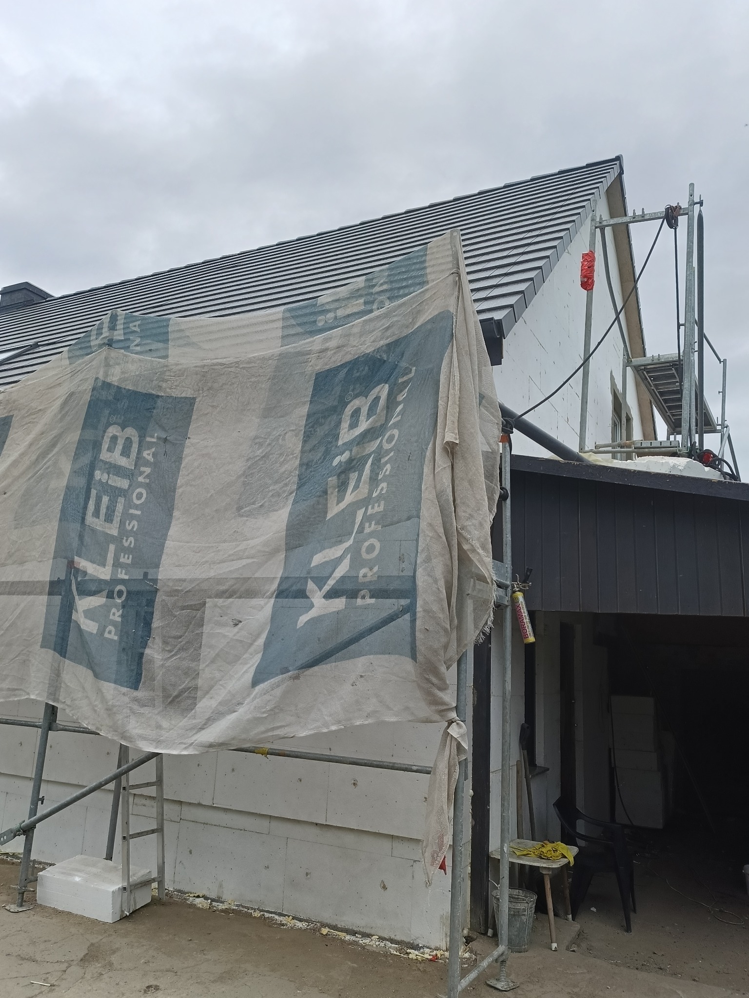 Dom w trakcie ocieplania, widoczne rusztowanie i siatka ochronna z logo Kleib Professional, szara dachówka, ściany z białego styropianu.