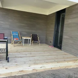 Drewniany taras z dwoma krzesłami i grillem. Jasne deski, poziome panele ścienne w odcieniach brązu. Widoczne schodki prowadzące na taras i element okna z żaluzjami.