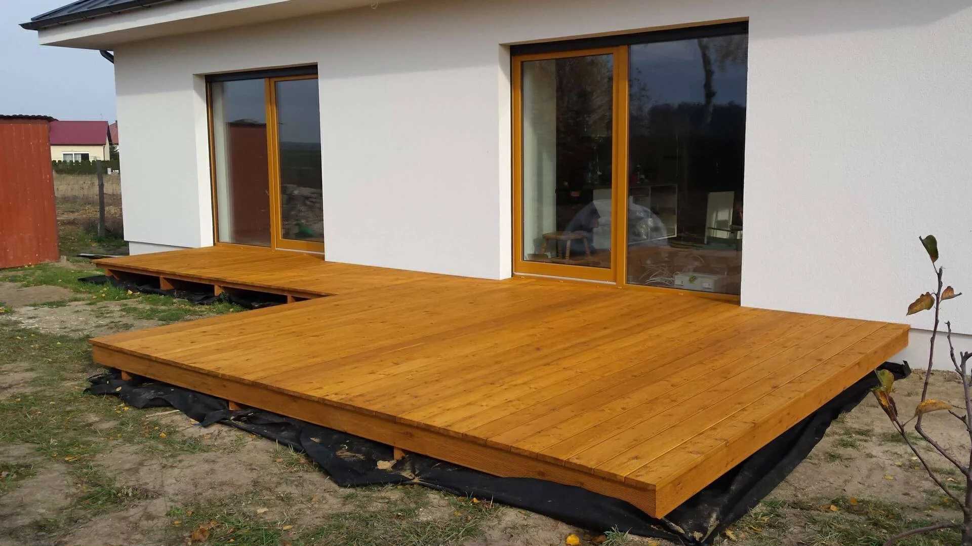 Nowoczesny taras z drewna w kształcie litery L, przylegający do białego budynku z dużymi oknami przesuwnymi. Ułożony na czarnej membranie, z widoczną konstrukcją nośną.