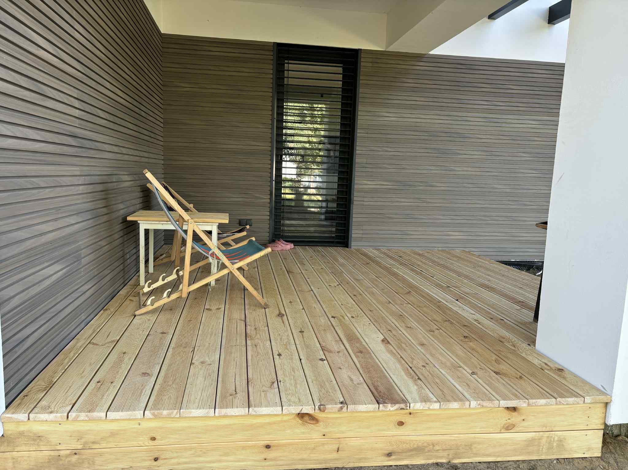 Jasny, drewniany taras z leżakiem i stolikiem na tle ściany z poziomych desek i wąskiego okna z żaluzjami. Widok na zewnątrz.