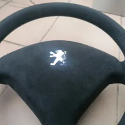 Obszyta czarną alcantarą kierownica Peugeot z logo lwa, widoczne szwy, leżąca na jasnej powierzchni. Detal tapicerki samochodowej po renowacji.