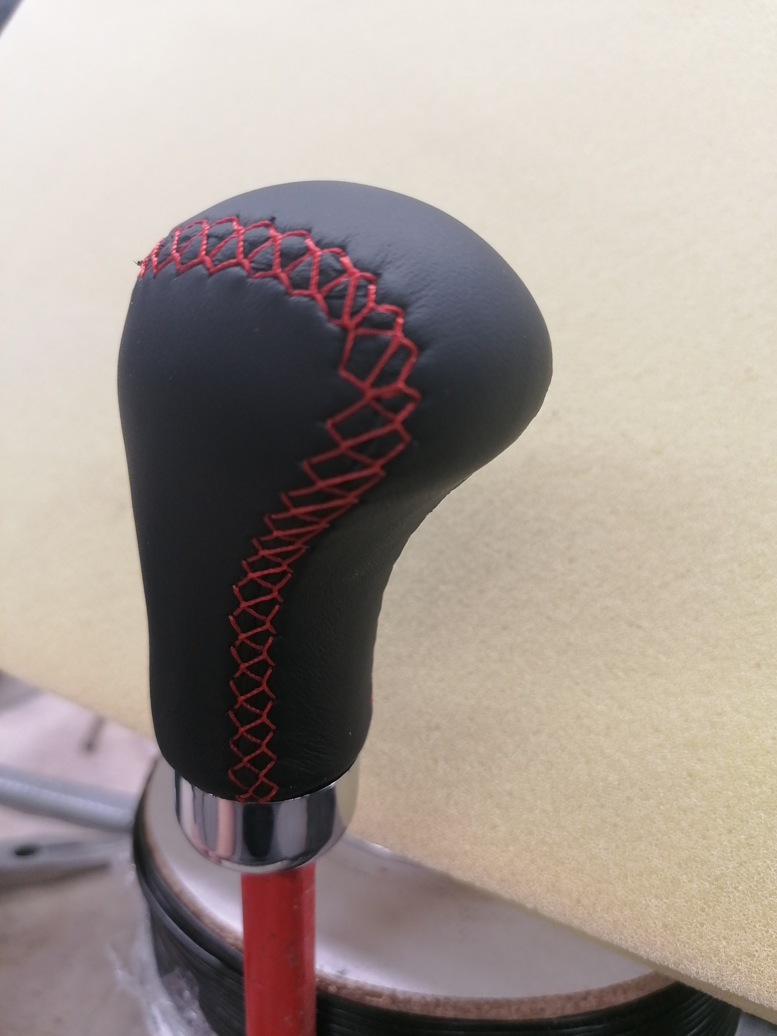 Czarna, skórzana gałka zmiany biegów z czerwonym szwem krzyżykowym, osadzona na metalowej podstawie i czerwonym pręcie, prezentowana na jasnym tle.