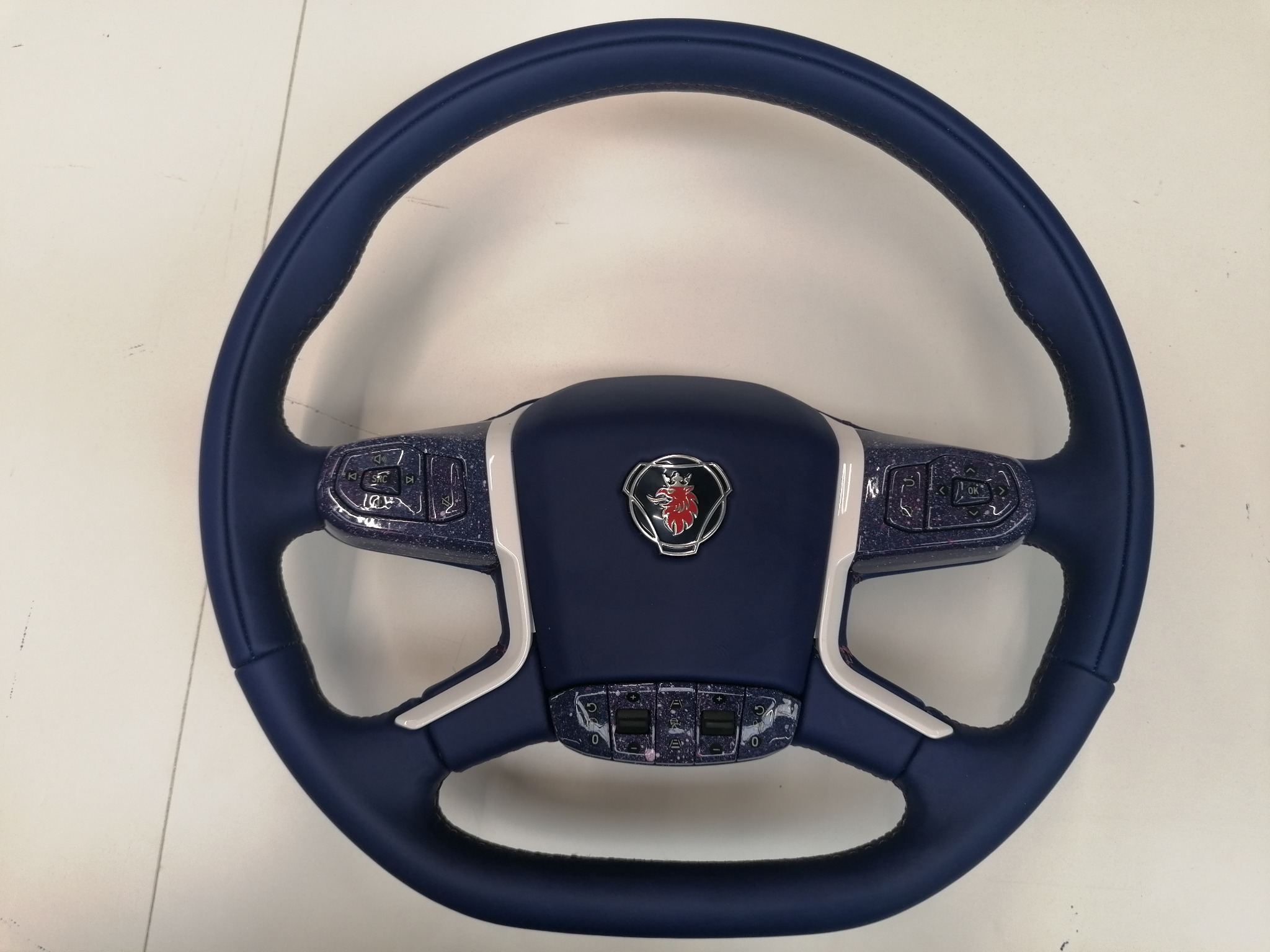 Granatowa, obszyta skórą kierownica Scania z panelami sterowania. Wykończenie z efektem brokatu. Chromowane logo na środku. Precyzyjne przeszycia.