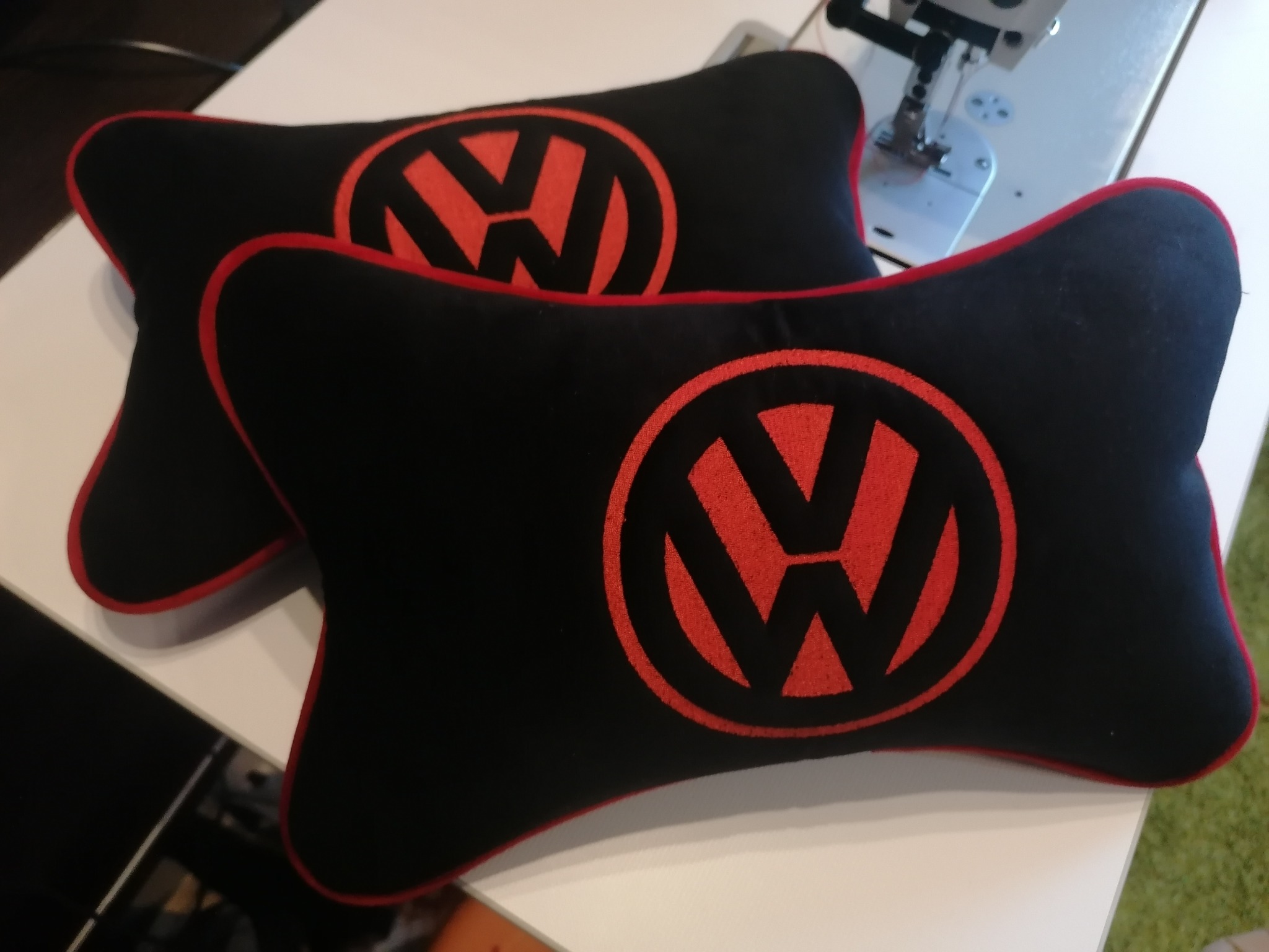 Dwie czarne poduszki samochodowe z czerwonym logo VW, leżące na białym stole obok maszyny do szycia. Poduszki mają kształt kości i czerwoną lamówkę.