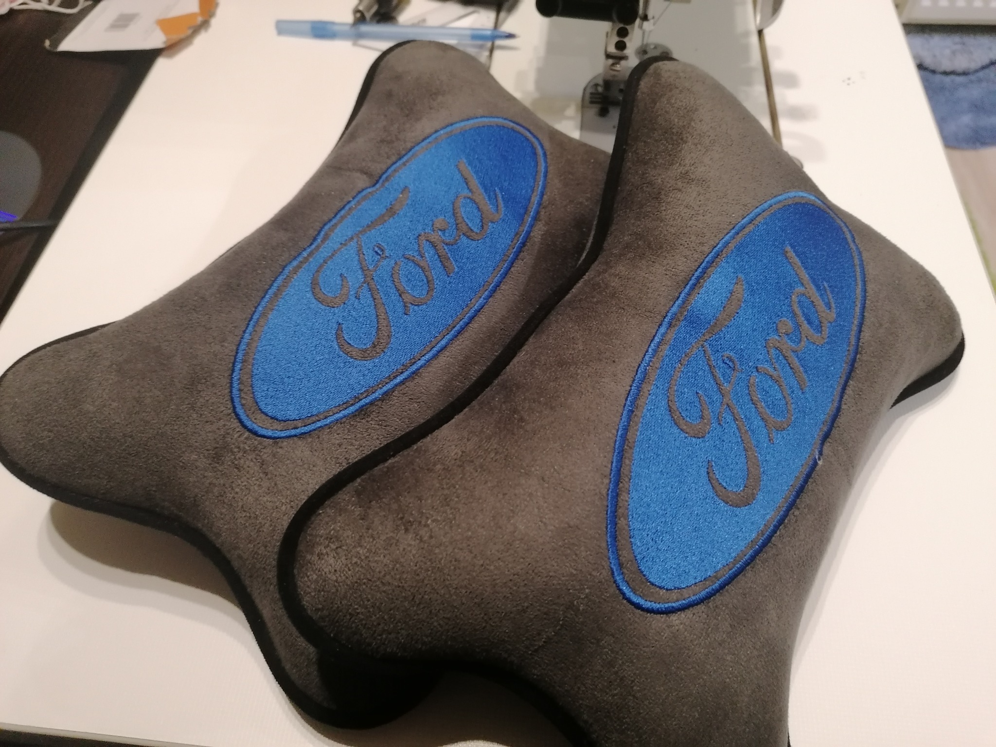 Dwie szare poduszki samochodowe z wyhaftowanym niebieskim logo 'Ford', leżące na białym stole, obok maszyny do szycia.