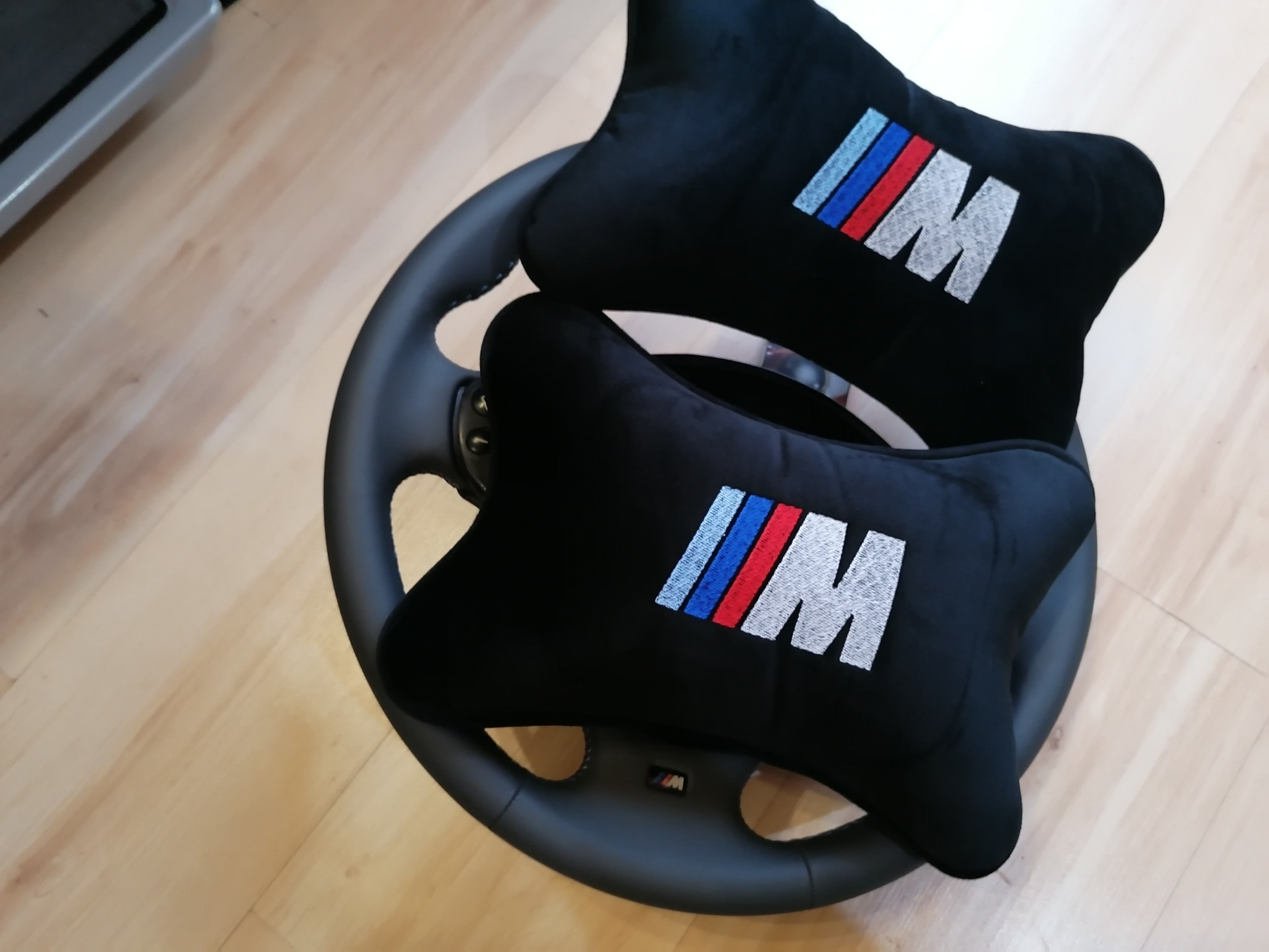 Dwa czarne zagłówki samochodowe z haftowanym logo BMW M na kierownicy. Drewniana podłoga w tle.